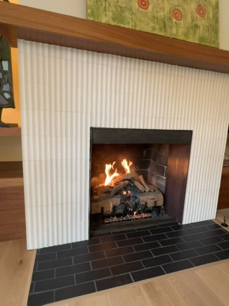 A fireplace with a burning fire inside, surrounded by a white tiled facade and a wooden mantel. The hearth area has black bricks and a wooden floor nearby.