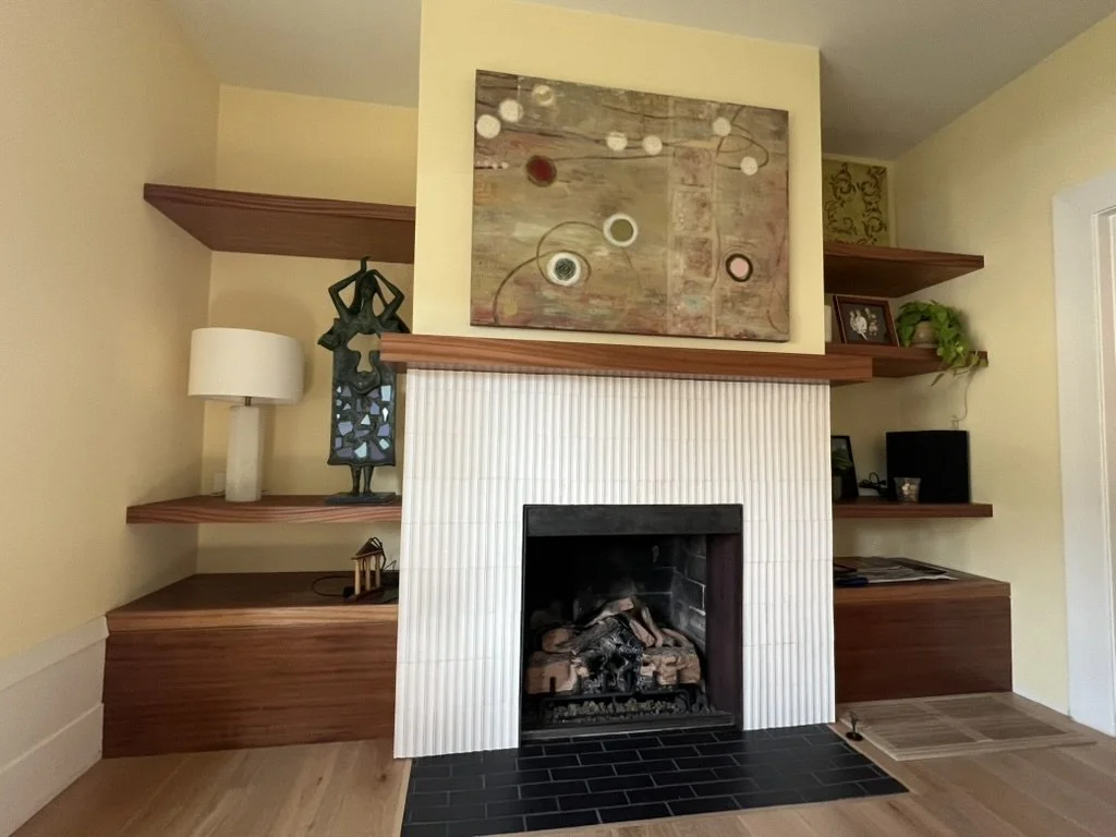 Living room with a fireplace surrounded by wooden shelves, decorative artwork above, a sculpture on the left shelf, a table lamp, a framed photo, a potted plant, and a small decorative piece on the right shelf.