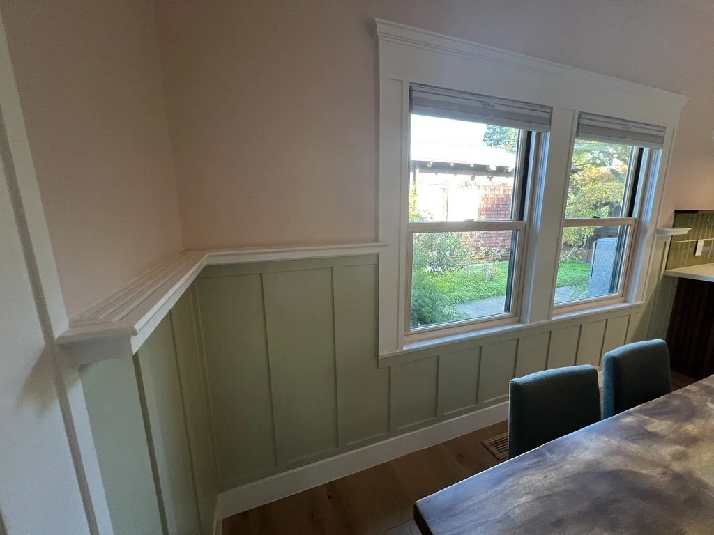 Interior of a dining area with beige walls, a window with white trim, and green wainscoting below the window. Part of a wooden dining table with two dark green chairs is visible.