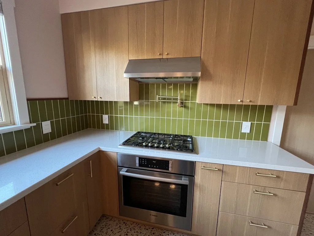 Kitchen with wooden cabinets, green tiled backsplash, white countertop, built-in gas stove and oven, and a window on the left.