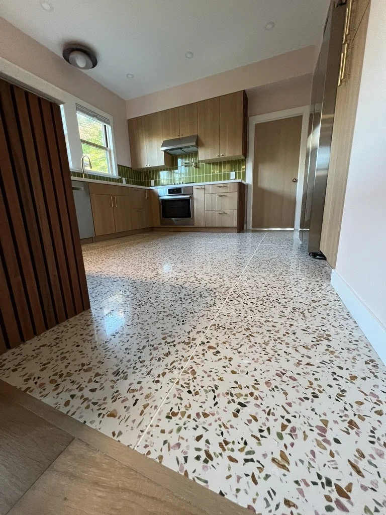 Modern kitchen with terrazzo floor, wood cabinets, green tile backsplash, stainless steel oven and refrigerator, and a window with a view of greenery.