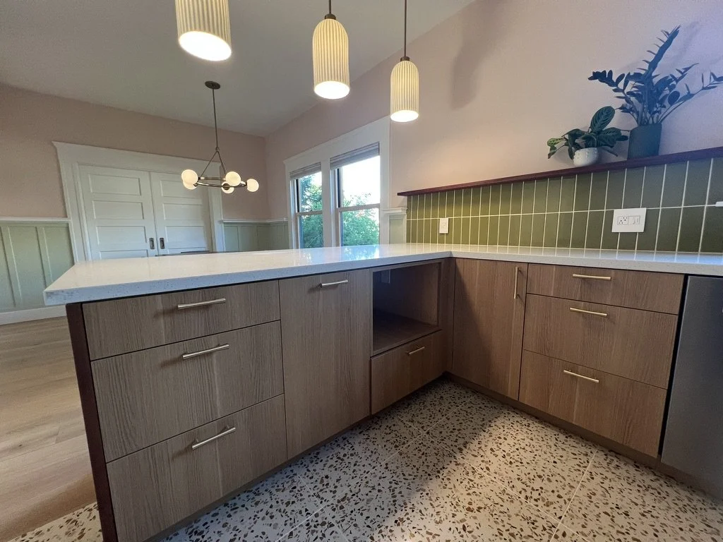 Modern kitchen with wooden cabinets, speckled floor tiles, green tiled backsplash, and white countertops. Potted plants on a raised shelf and pendant lights hanging from the ceiling.