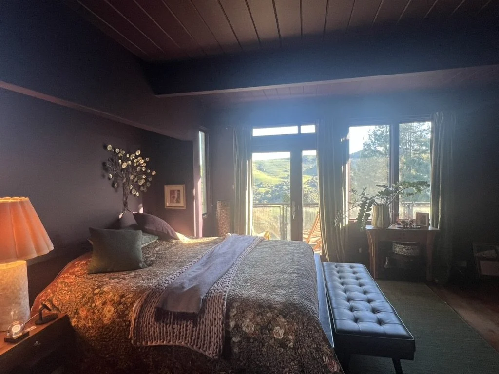 A cozy bedroom with a metal wall art above the bed, a nightstand with a lamp, a quilted bedspread, a bench at the foot of the bed, and a balcony door with greenery outside.