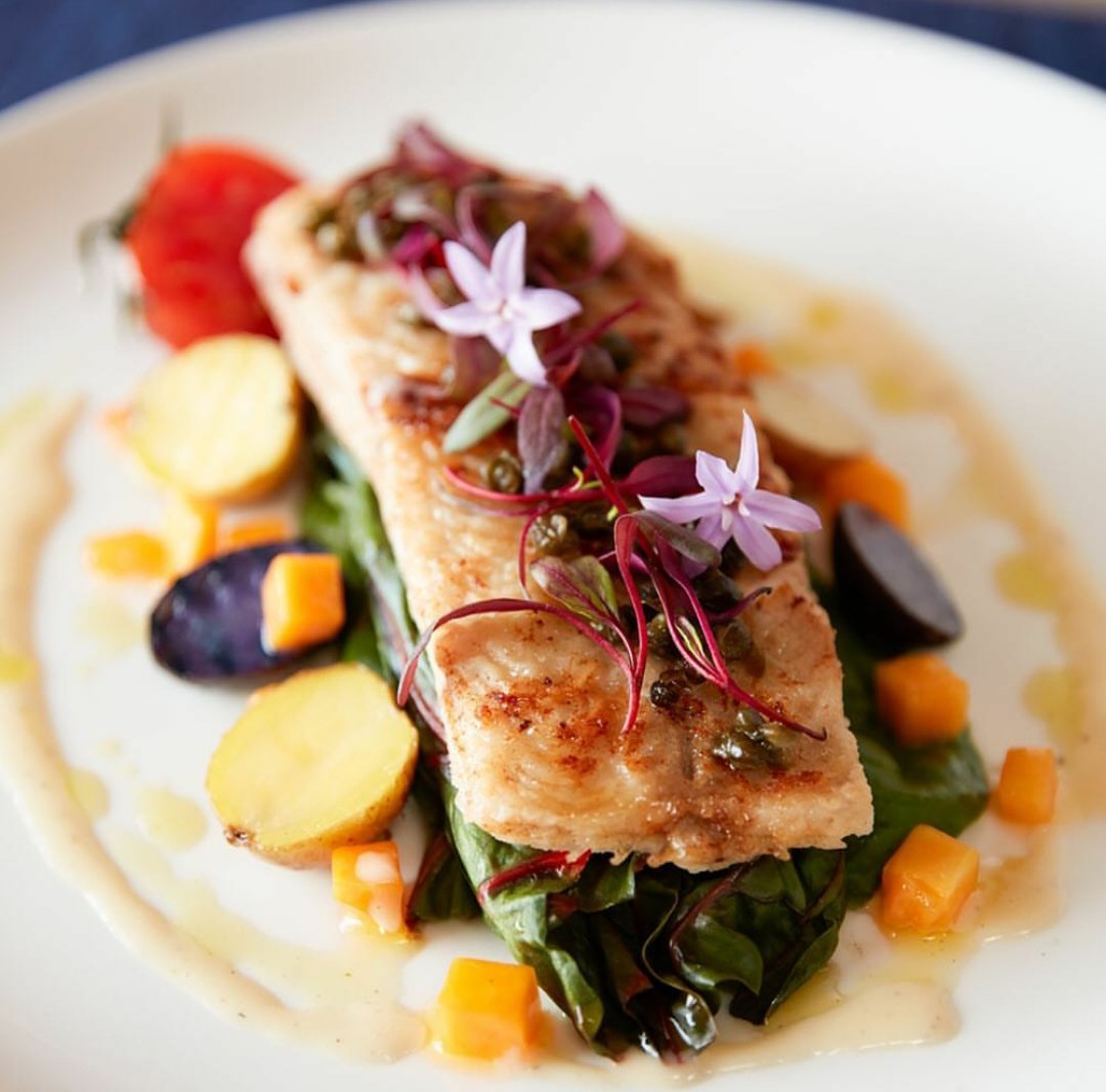 Pan-seared fish fillet garnished with edible flowers, served over mixed vegetables including potatoes, carrots, olives, and greens with a light sauce.