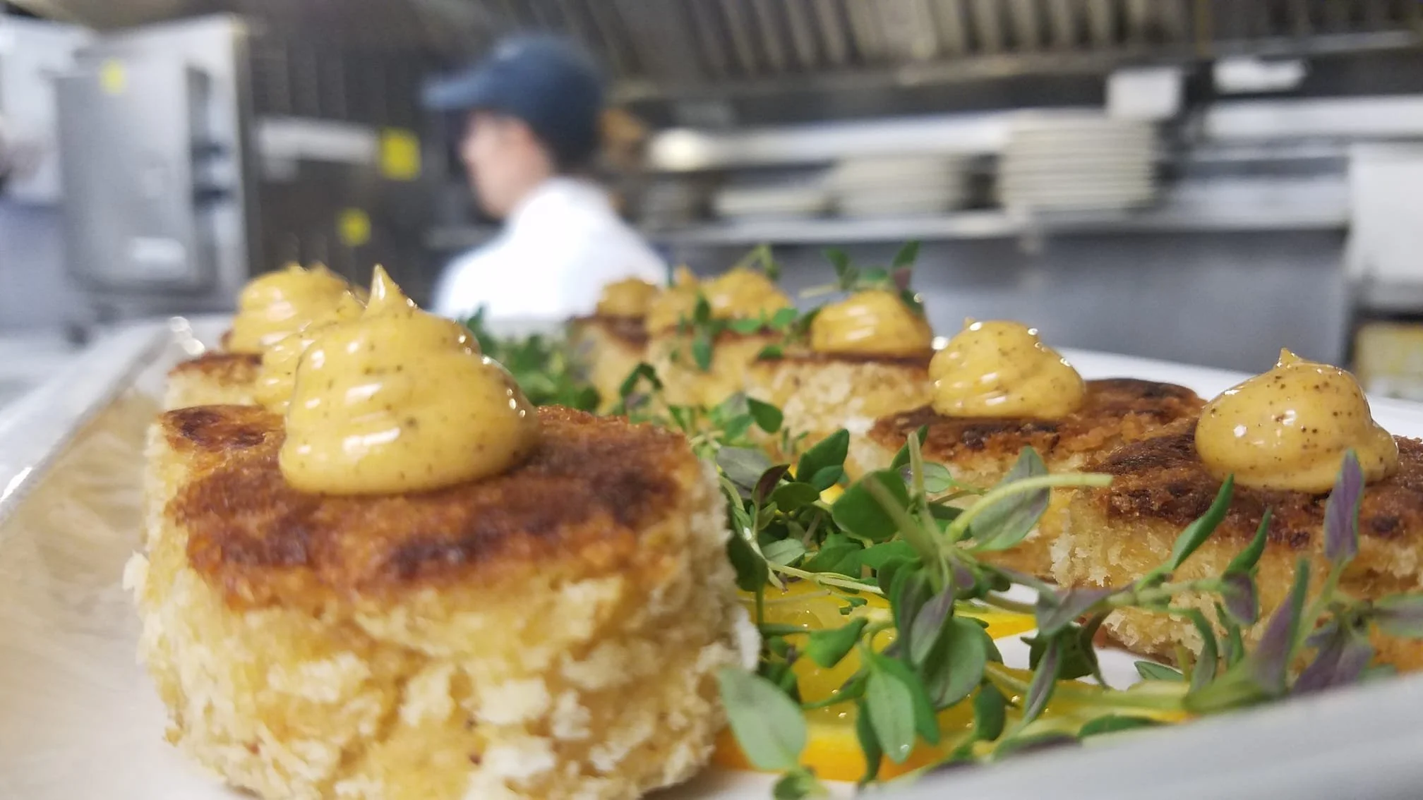Close-up of plated gourmet dishes with a yellow sauce dollop, garnished with microgreens, set in a professional kitchen with a chef in the background.