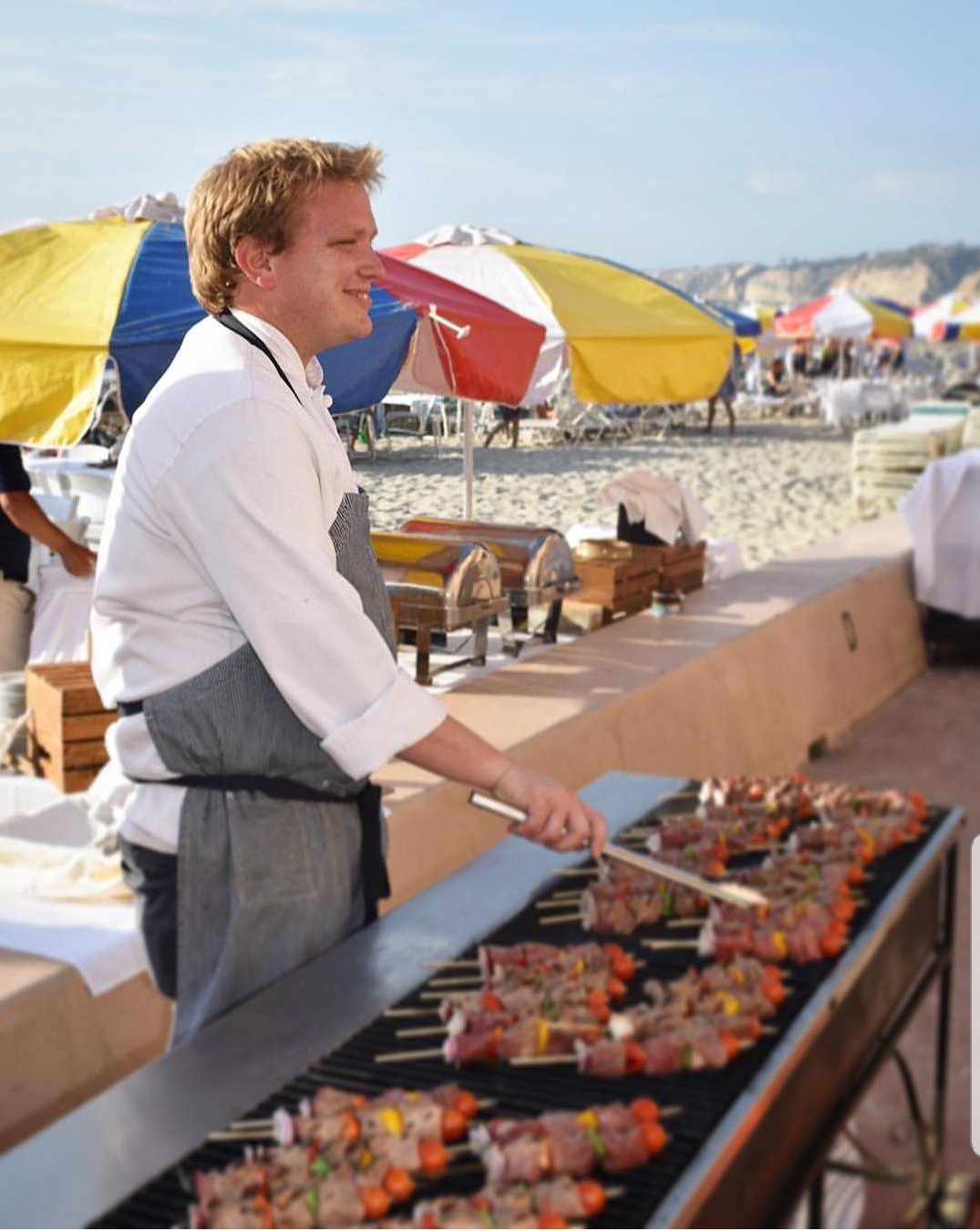 A man grilling skewered meat on a barbecue at the beach with colorful umbrellas and people in the background.