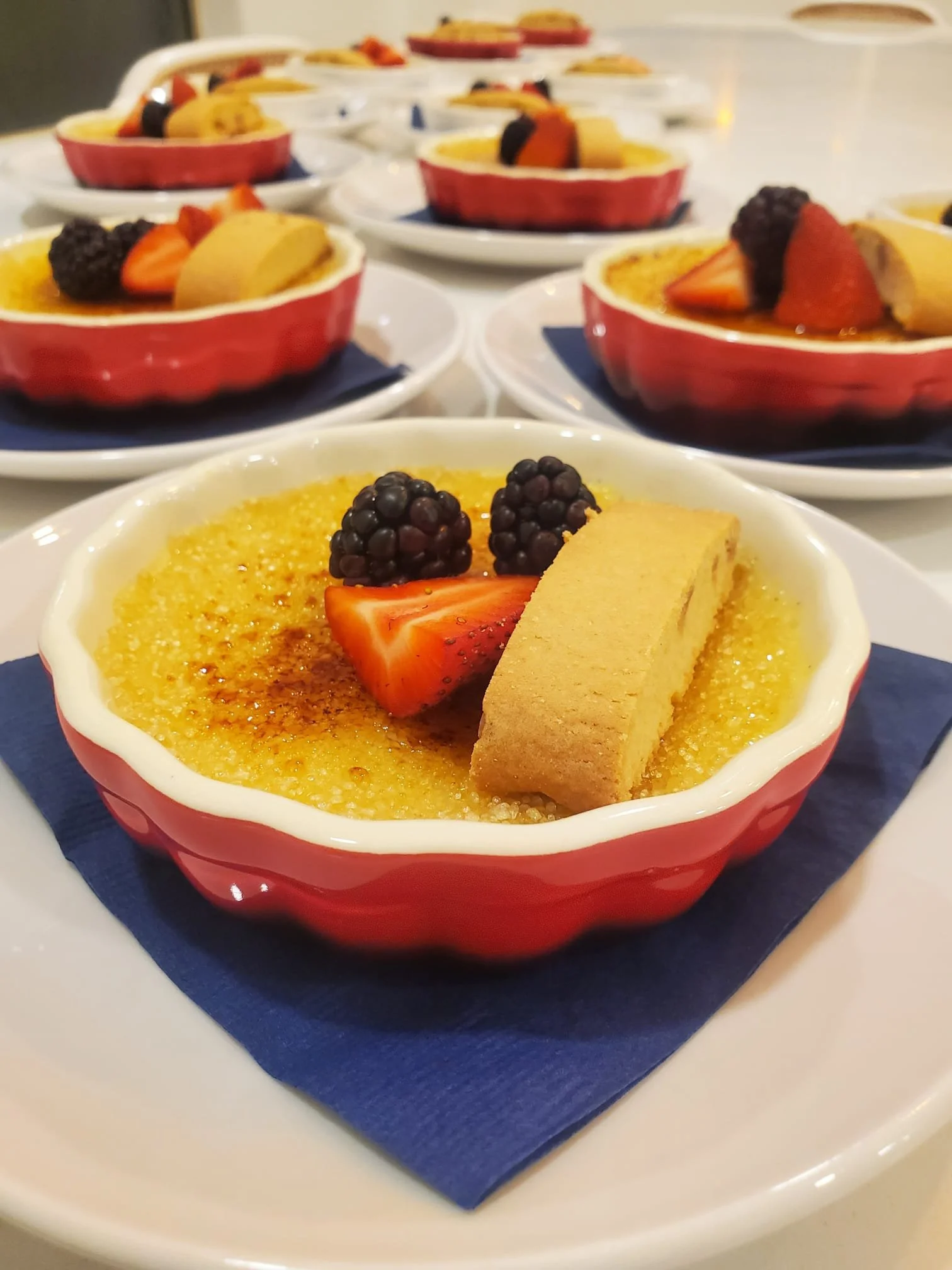 Creamy crème brûlée topped with strawberries, blackberries, and a shortbread cookie, served in a red ramekin on a blue napkin.