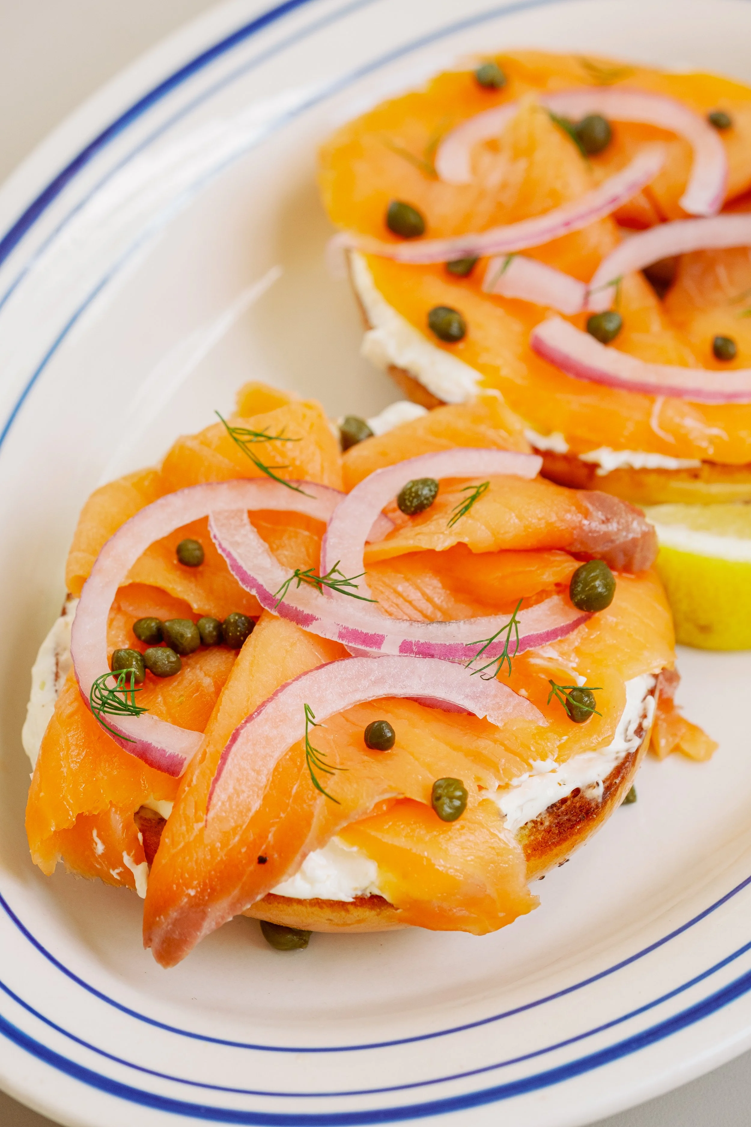 Smoked salmon on bagel with cream cheese, red onion slices, capers, and dill, served with lemon wedge on a white plate with blue stripes.
