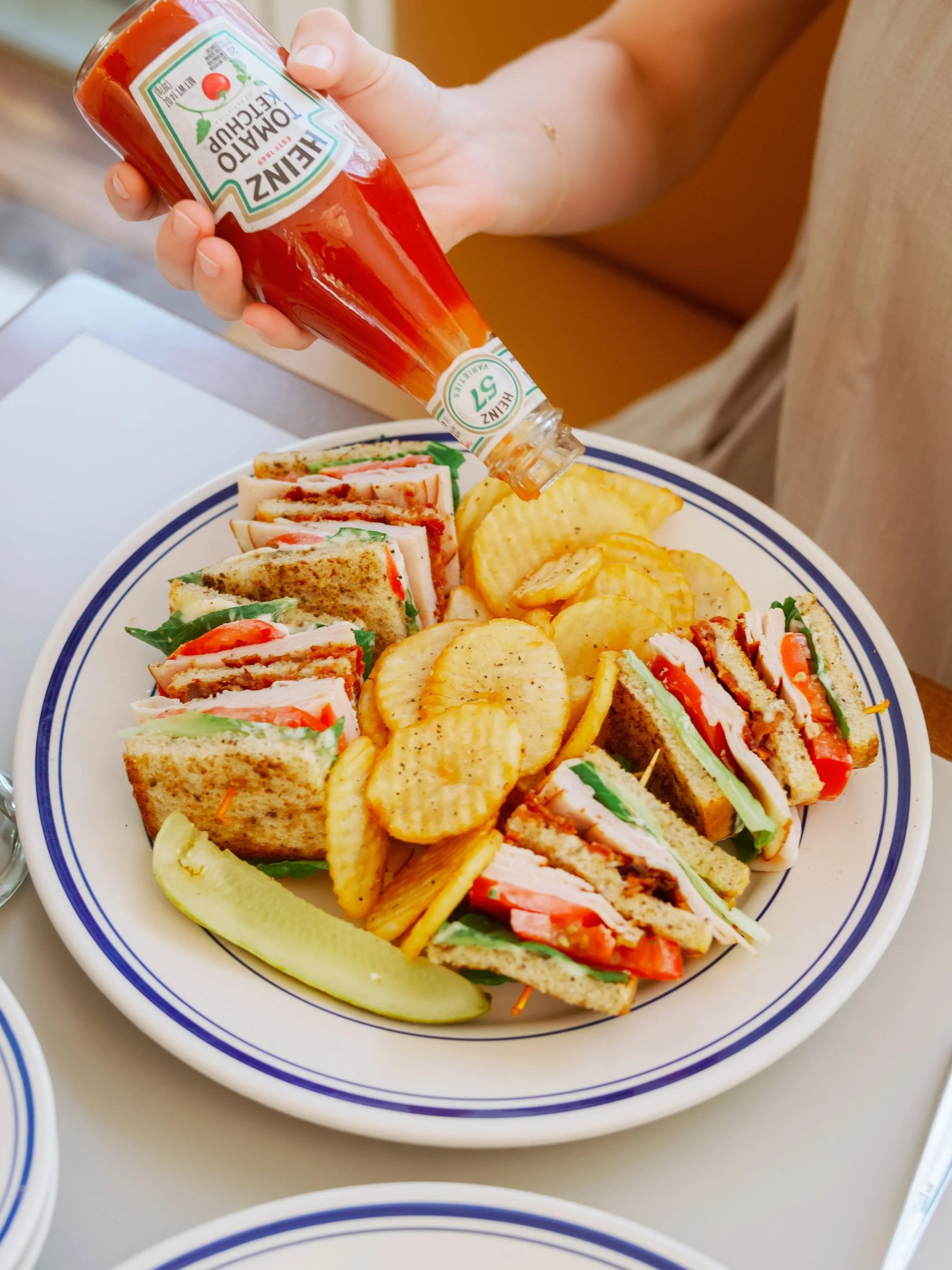 A white plate with a blue rim holding club sandwiches, potato chips, and pickle slices. A person is squeezing Heinz tomato ketchup onto the sandwich.
