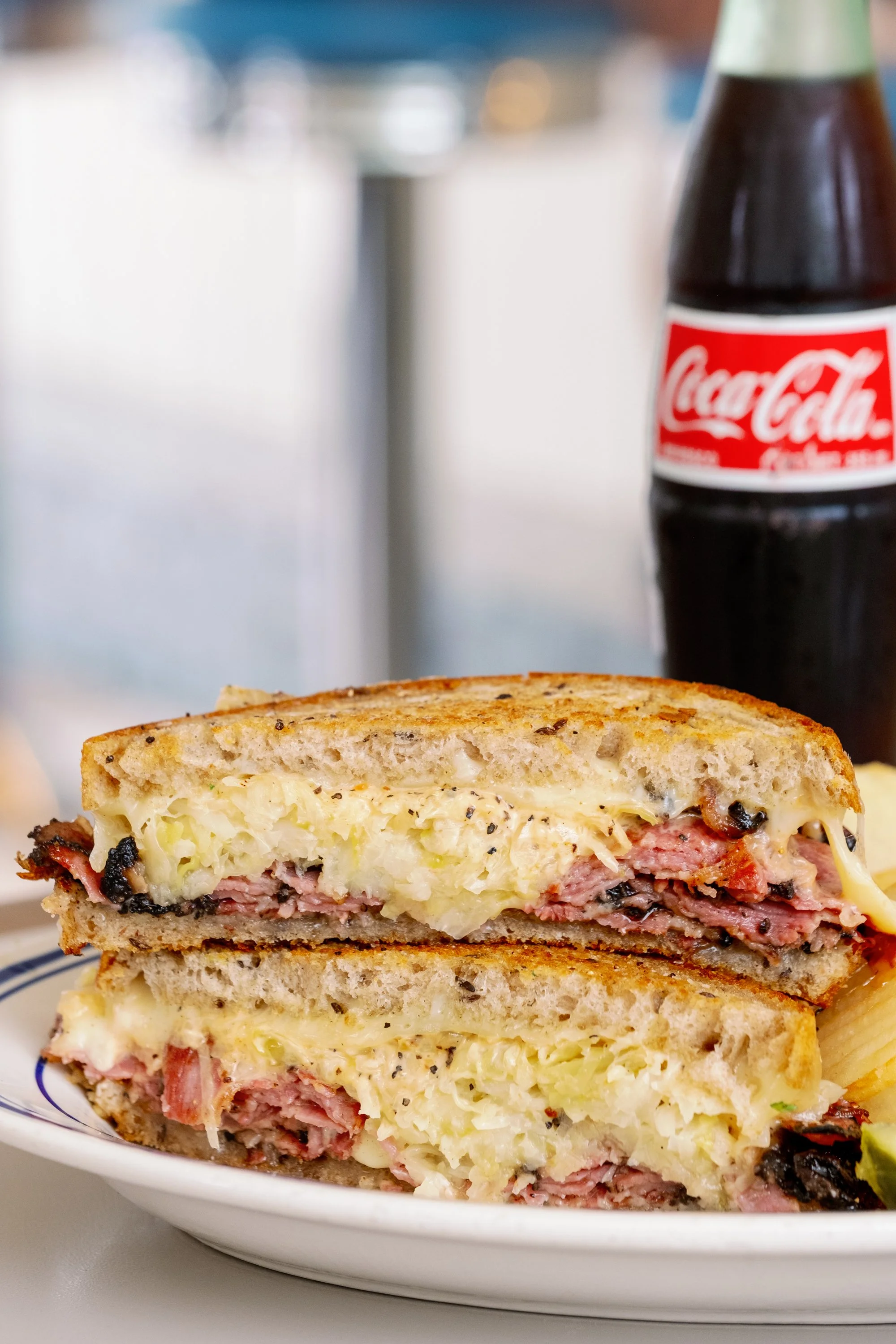 Half of a toasted Reuben sandwich on a white plate with shredded cheese and potato chips, a Coca-Cola bottle in the background.