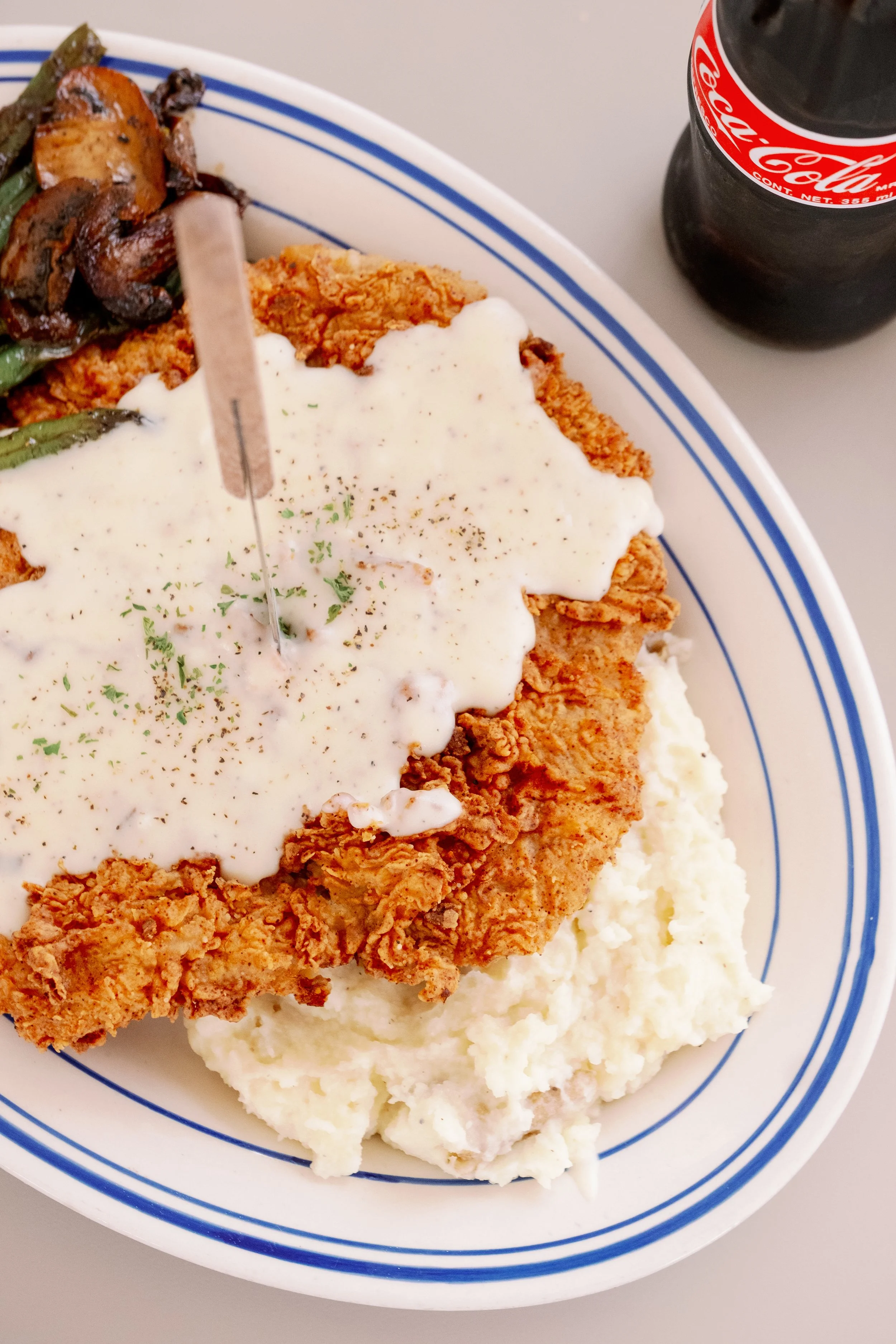 Plate of fried chicken with gravy, mashed potatoes, and green beans, with a small bottle of Coca-Cola in the background.