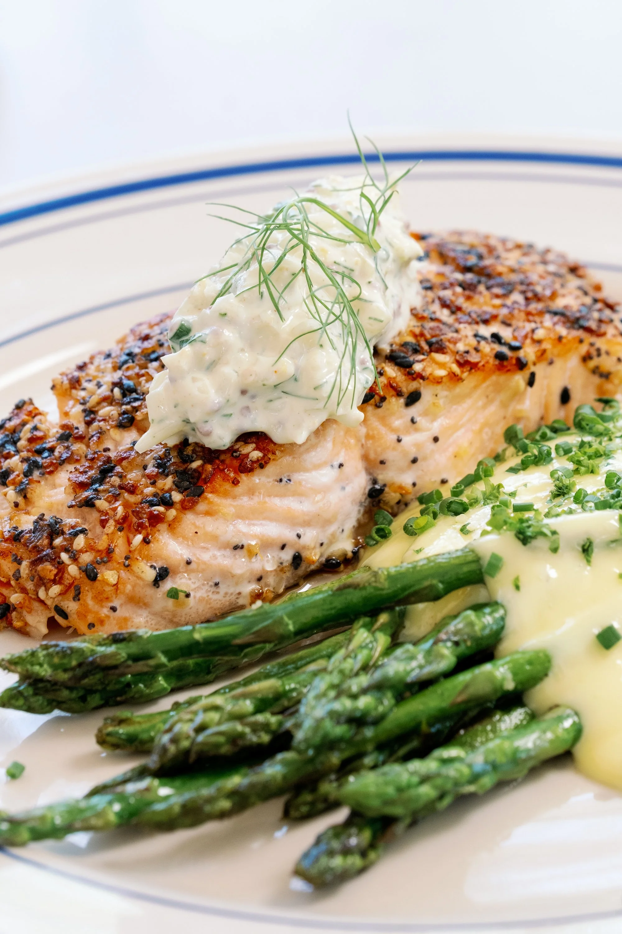 A plate of baked salmon topped with tartar sauce and herbs, served with roasted asparagus and a side of creamy mashed potatoes garnished with chives.