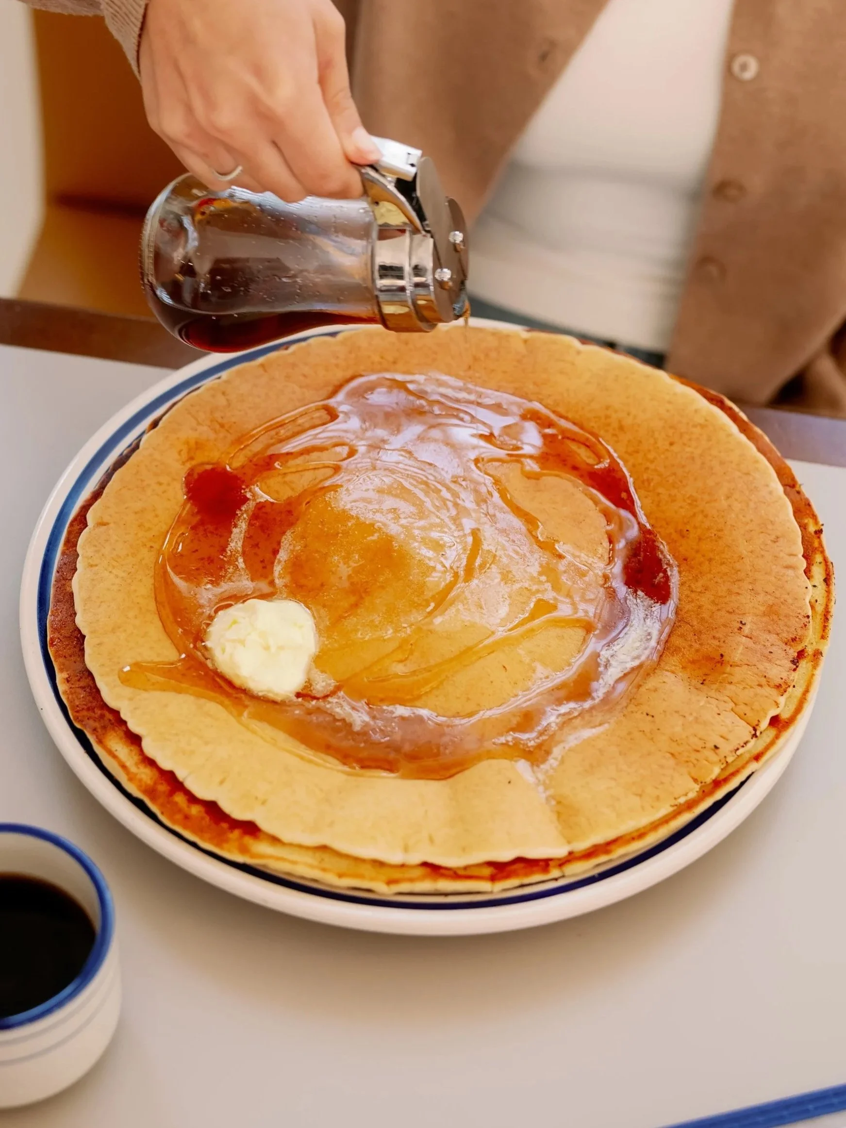 Person pouring syrup over a crepe topped with butter on a plate.