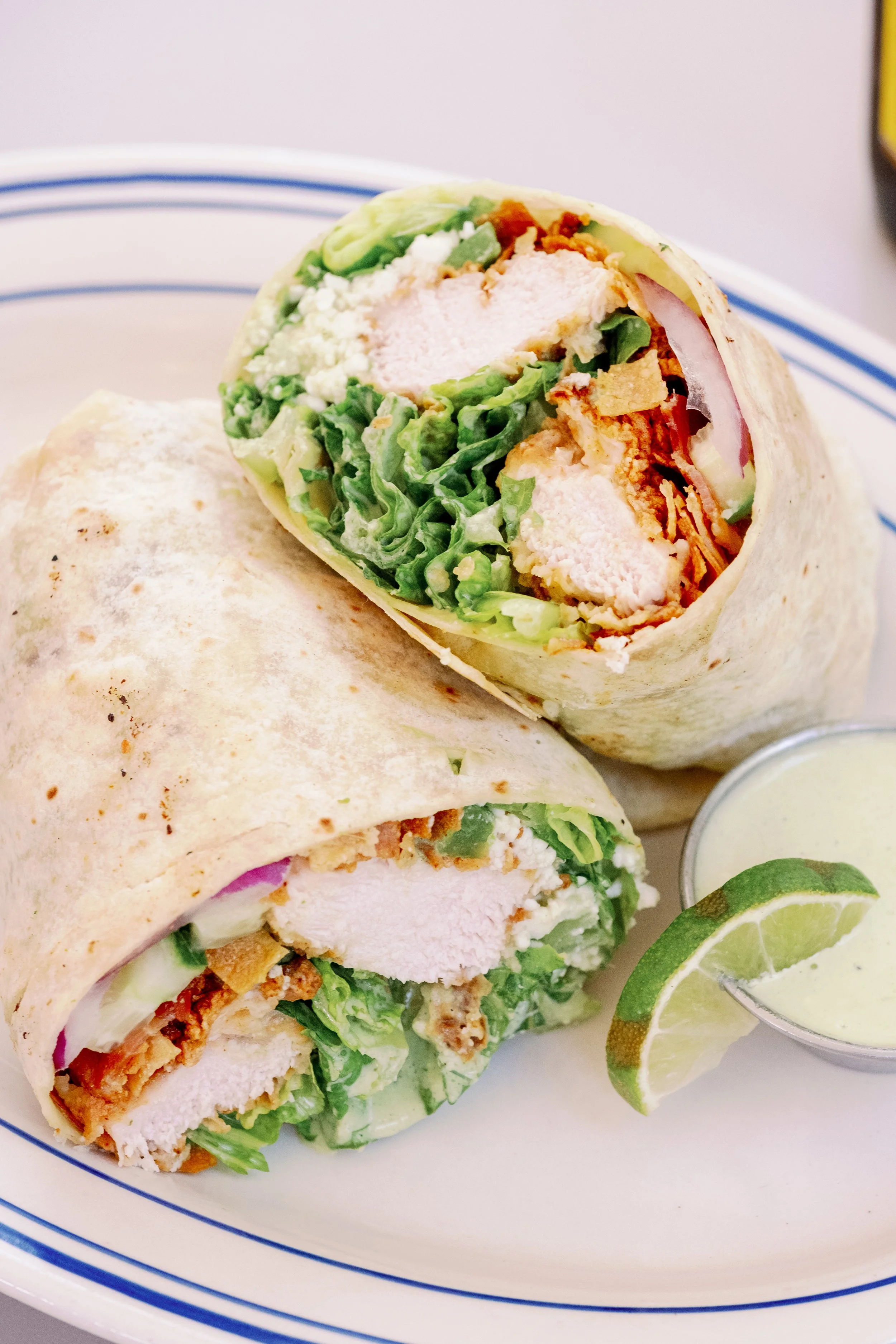 A plate with two chicken wrap sandwiches filled with lettuce, chicken, and vegetables, accompanied by a lime wedge and a small cup of green dipping sauce.