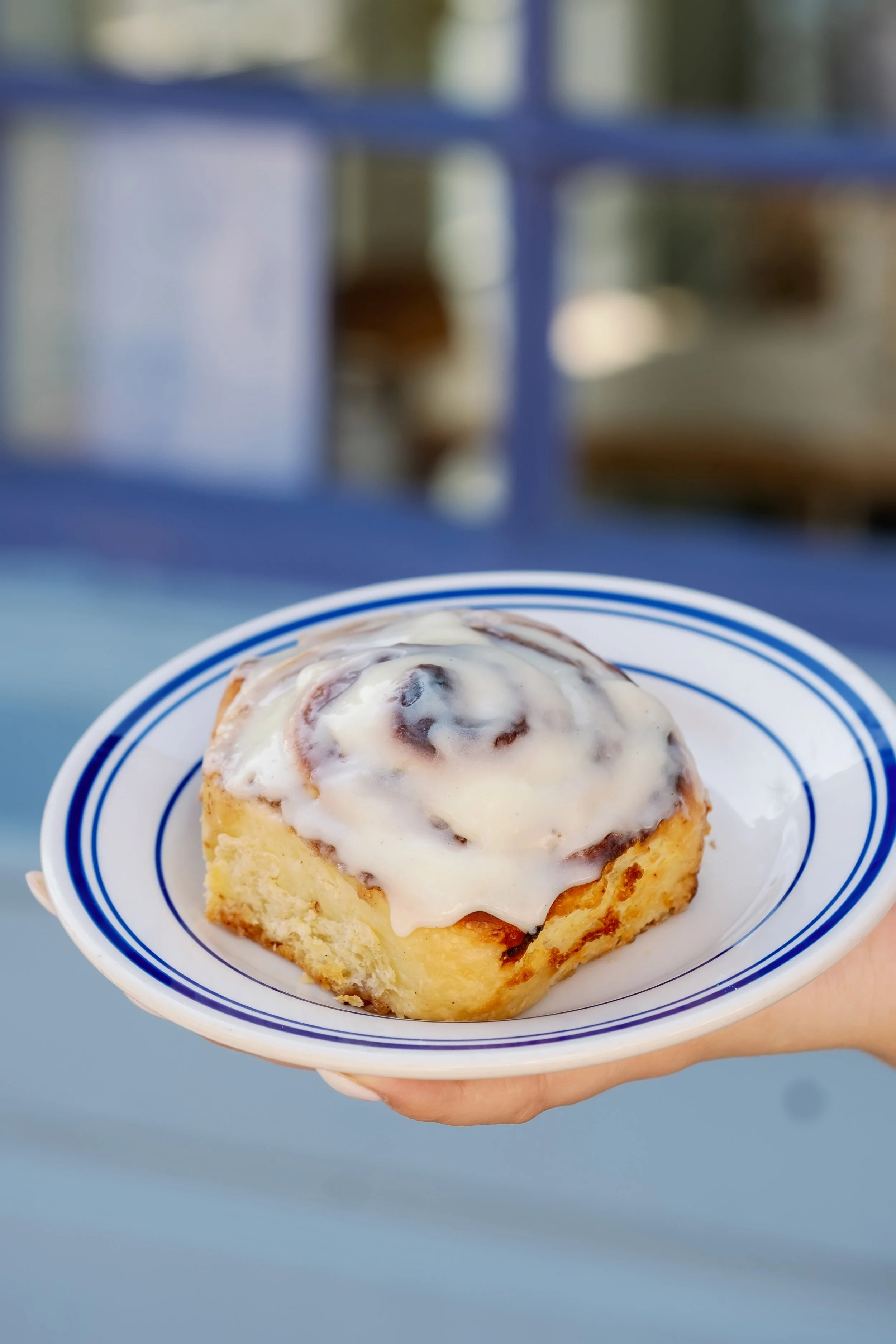 A cinnamon roll topped with white icing on a blue and white striped plate held by a person's hand.