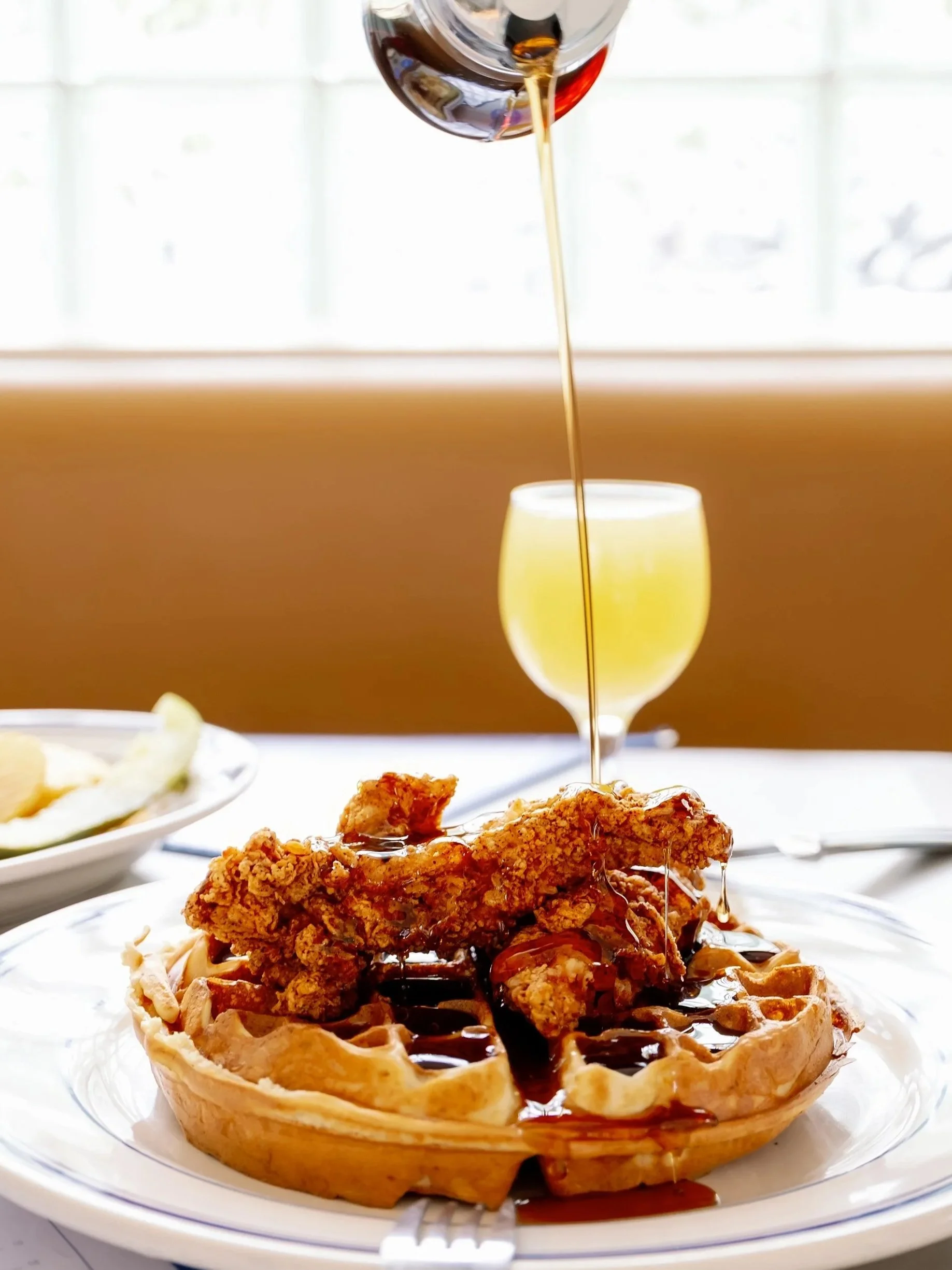 A waffle topped with fried chicken and drizzled with syrup, with a glass of wine and another beverage in the background.