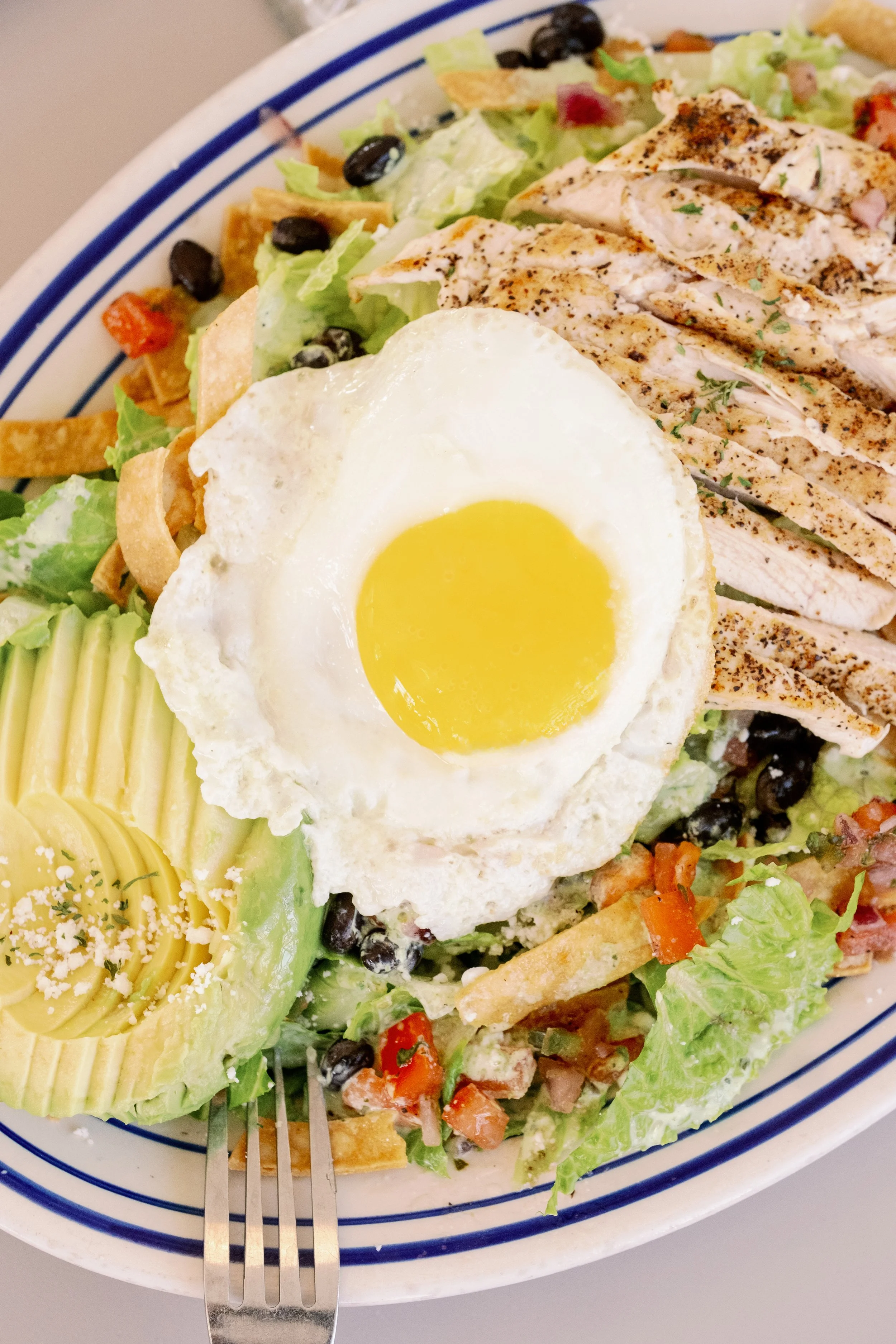 Lettuce salad topped with a fried egg, sliced grilled chicken, avocado, cherry tomatoes, black beans, tortilla strips, and shredded cheese in a bowl.