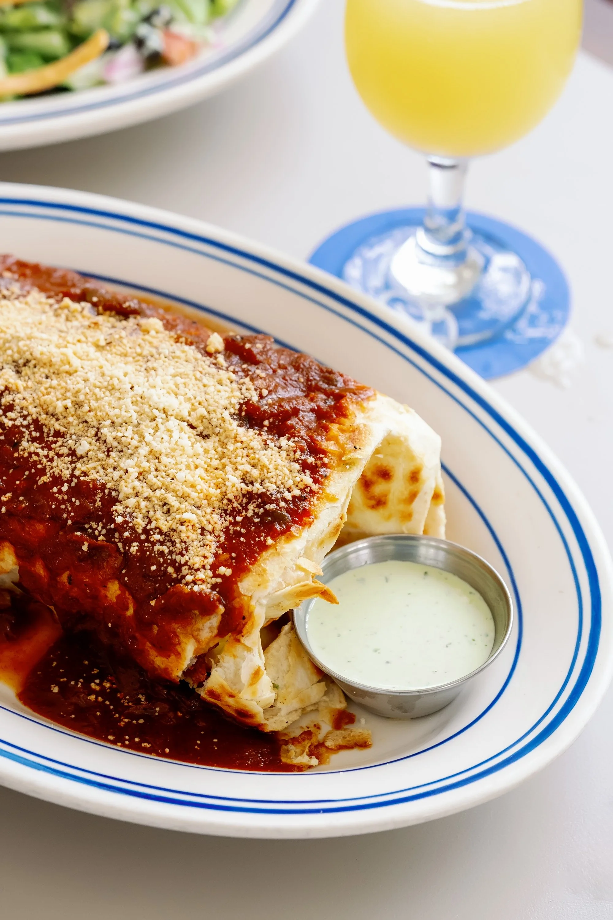 A plate of tamales topped with red sauce and grated cheese, served with a side of green sauce, and a glass of yellow beverage in the background.