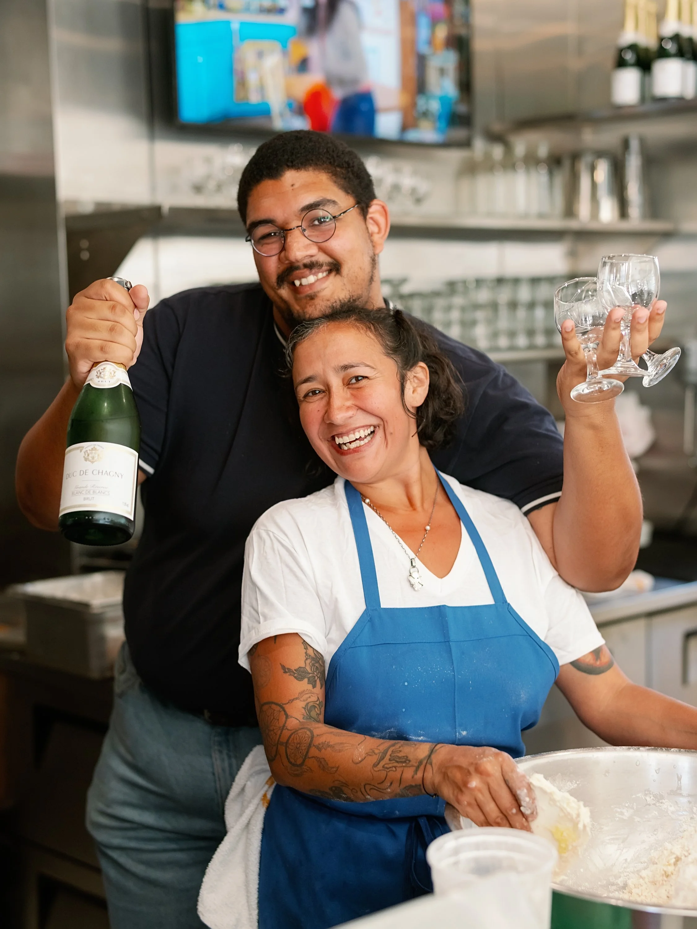 A man and woman smiling joyfully in a kitchen setting, celebrating with drinks; the man holds a bottle of champagne, and the woman holds three glasses. The woman is wearing a blue apron and has tattoos on her arms.