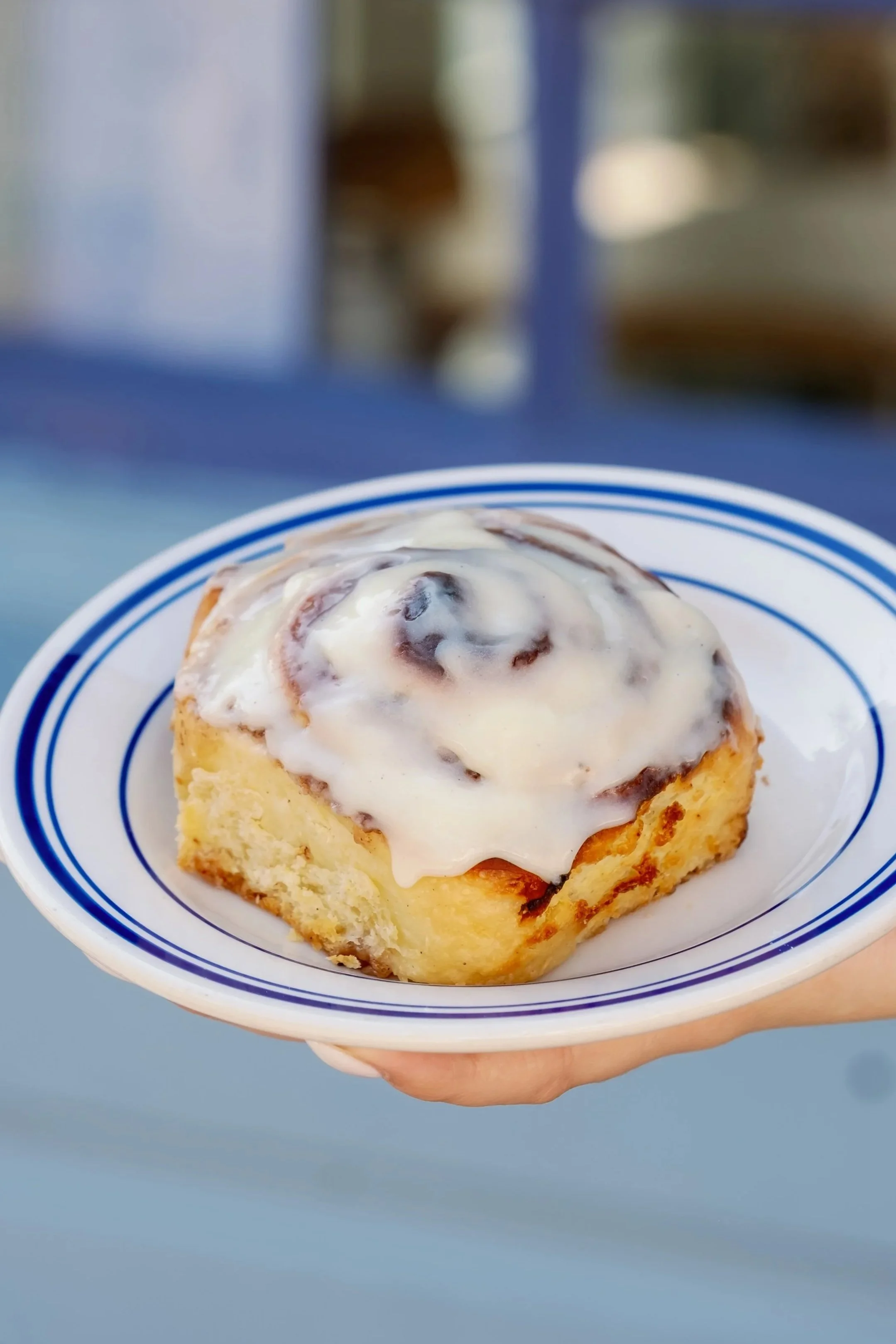 A cinnamon roll with icing on a white plate with blue stripes, held in a person's hand.