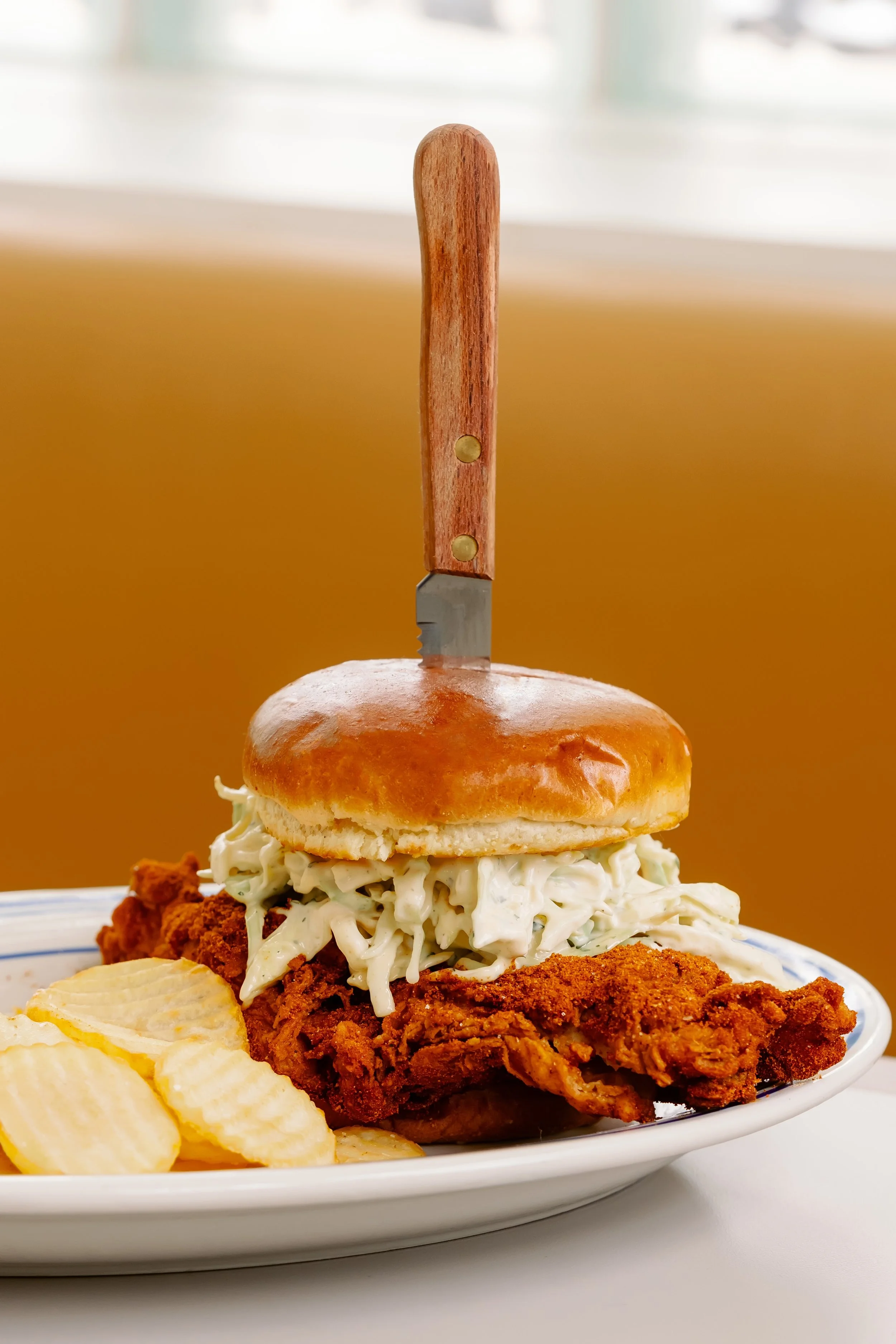 A plate with fried chicken, creamy coleslaw, potato chips, and a sandwich bun with a knife stabbed through the top.