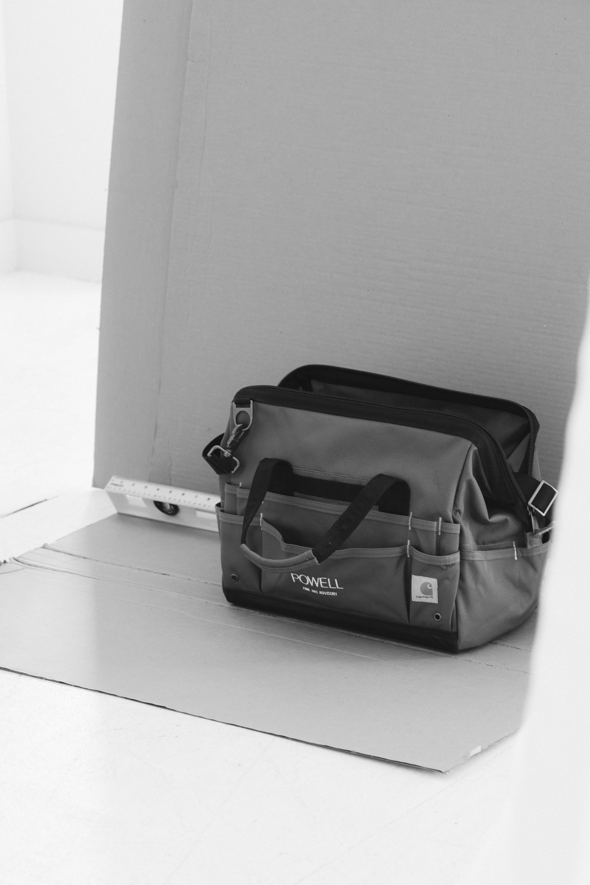 A gray and black Carhartt toolkit bag resting on a piece of cardboard on the floor, with a small level seen behind it.