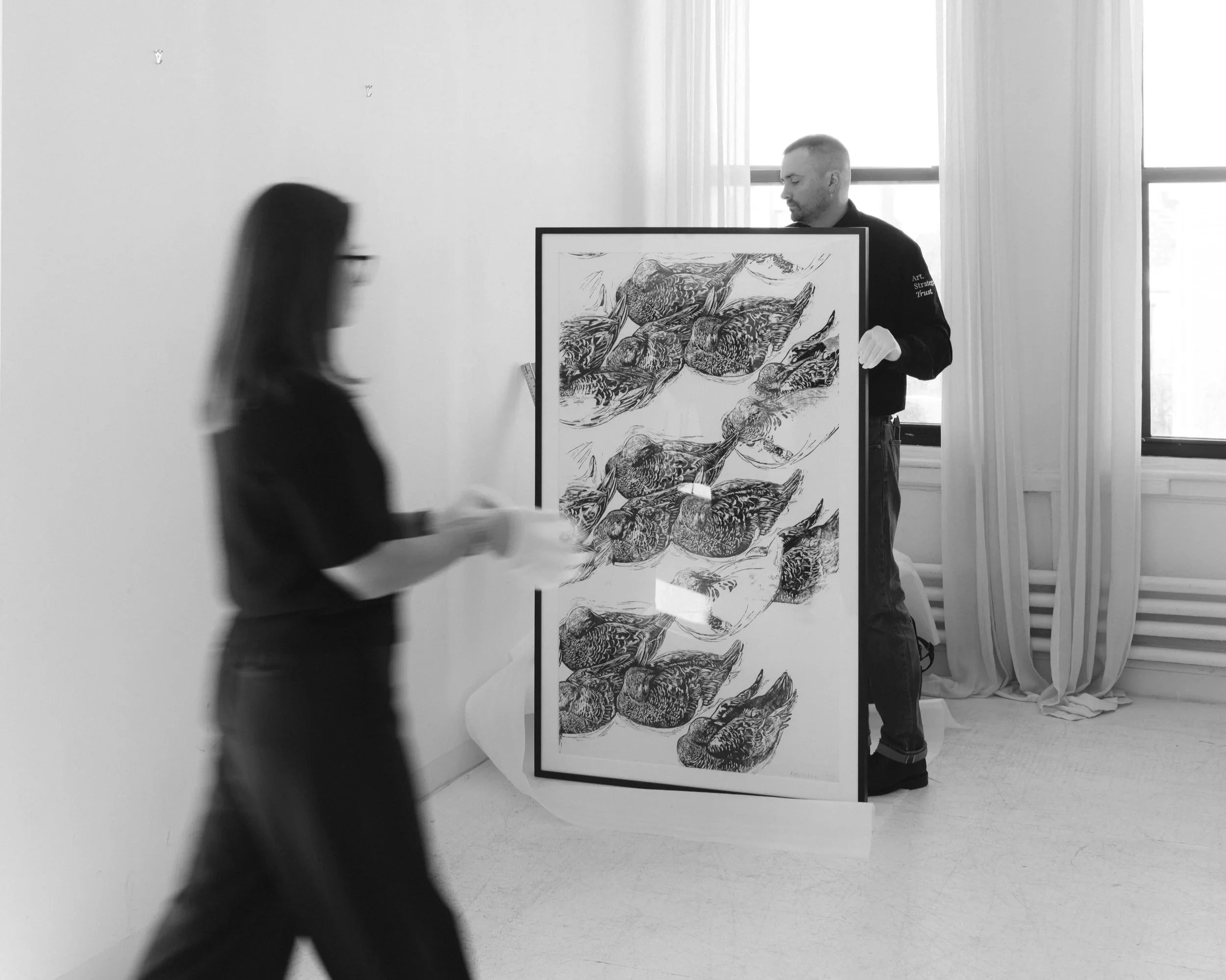 A man is holding a framed artwork while a woman walks past in a bright room with large windows and white curtains.