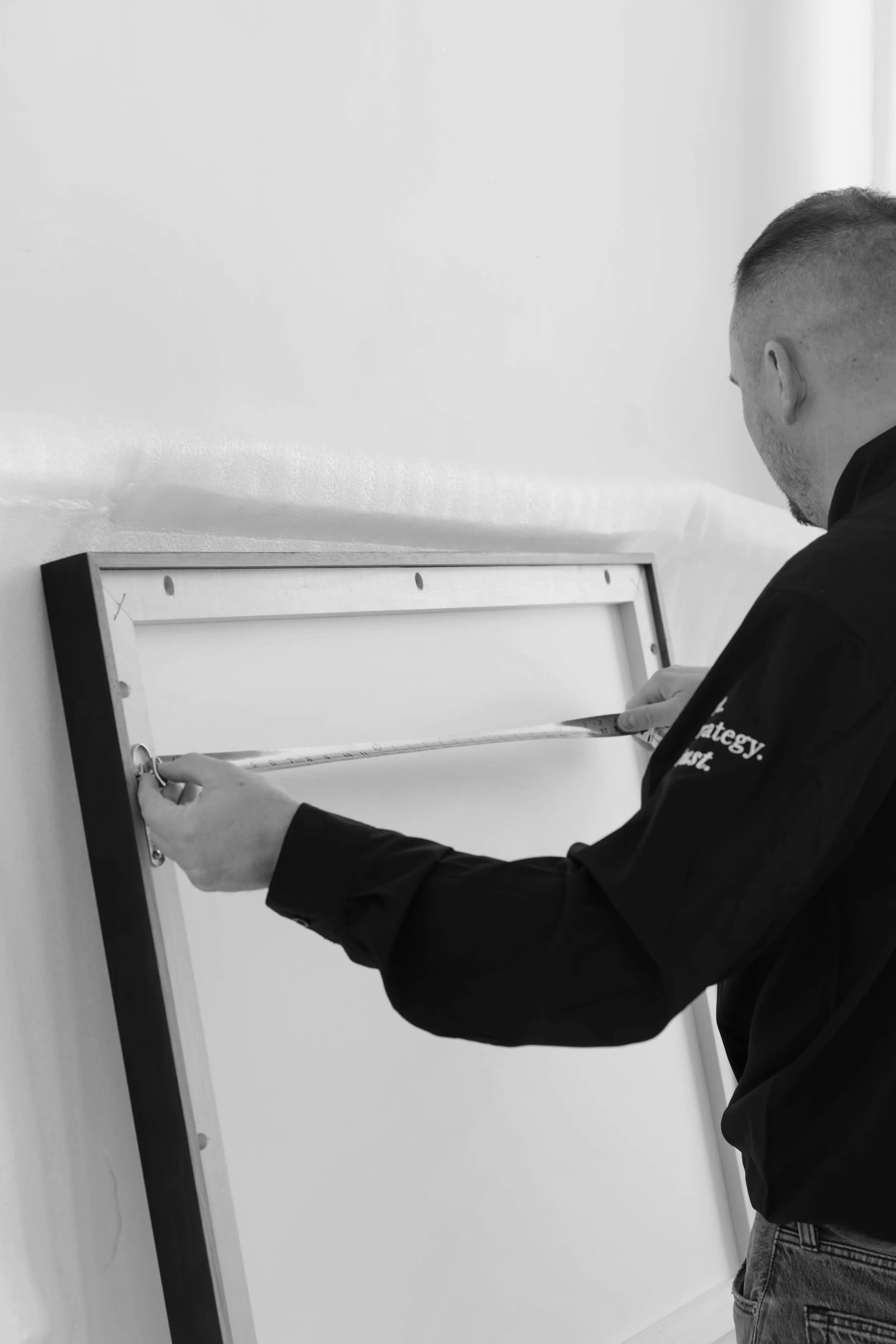 A man measuring a whiteboard with a measuring tape, standing by a wall.
