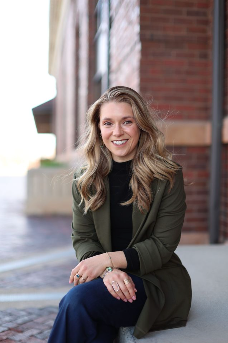 A woman with blonde hair, smiling, sitting on a concrete ledge outdoors near a brick building, wearing a black top, green blazer, and navy pants, with jewelry on her fingers and wrist.