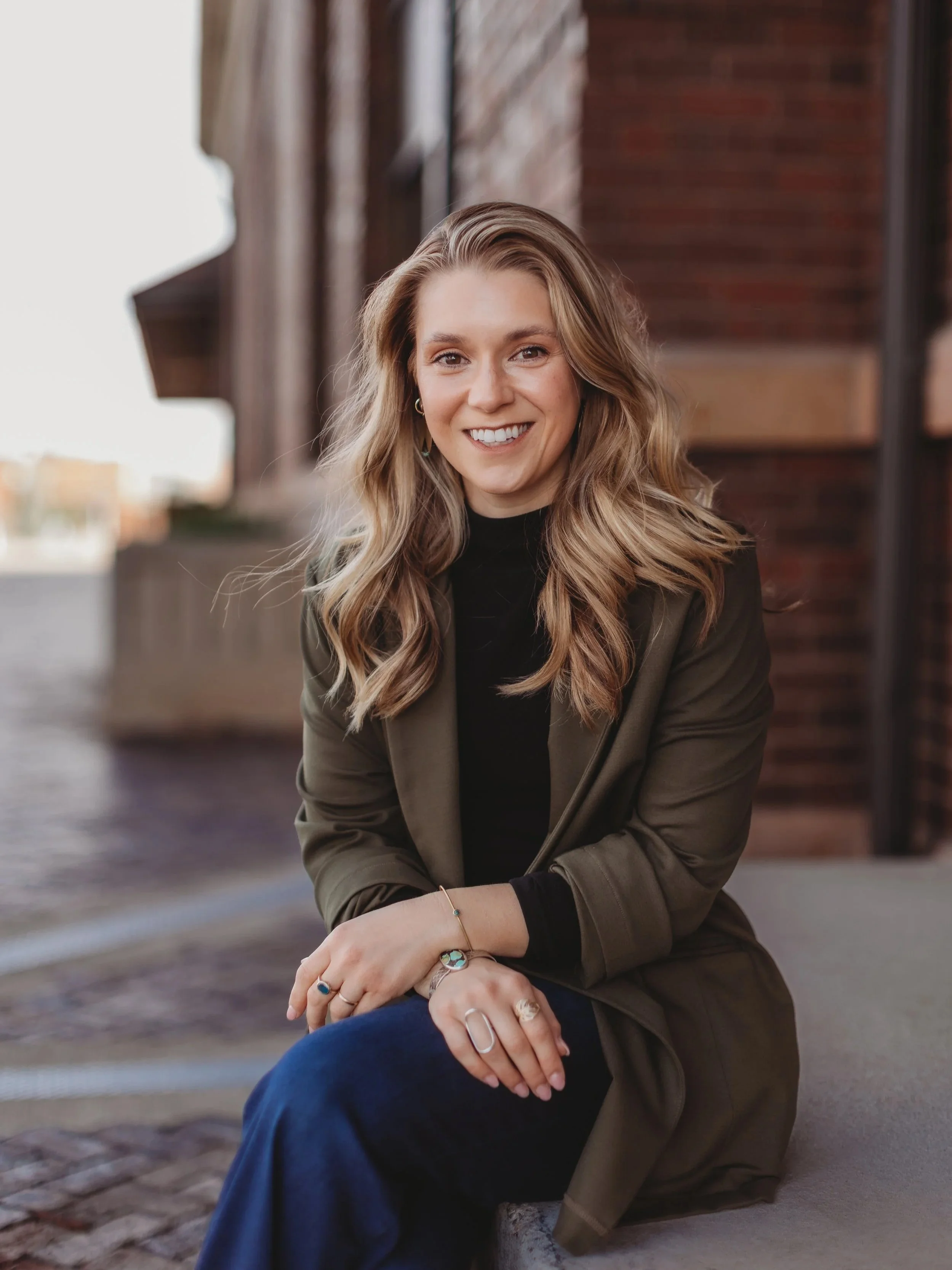 A smiling woman with wavy blonde hair, wearing a dark green blazer, black top, and jeans, sitting on a sidewalk against a brick wall with blurred city background.