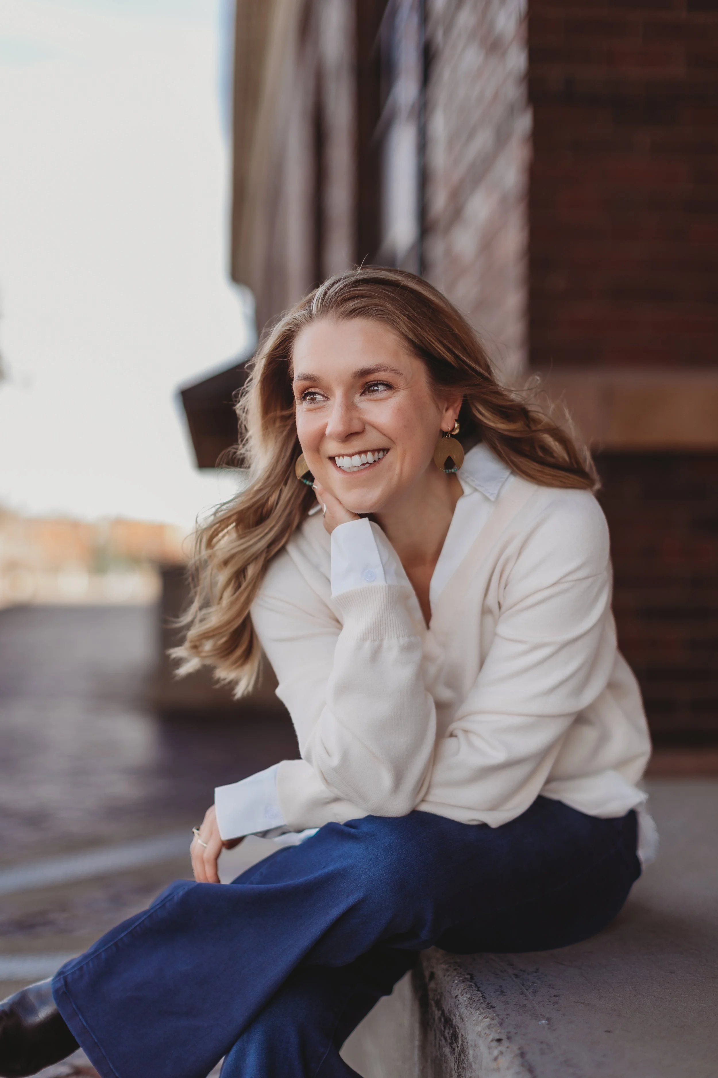 A young woman with long wavy hair, wearing a white shirt and dark blue pants, sitting on a stone ledge outdoors, smiling, with a wooden building in the background.