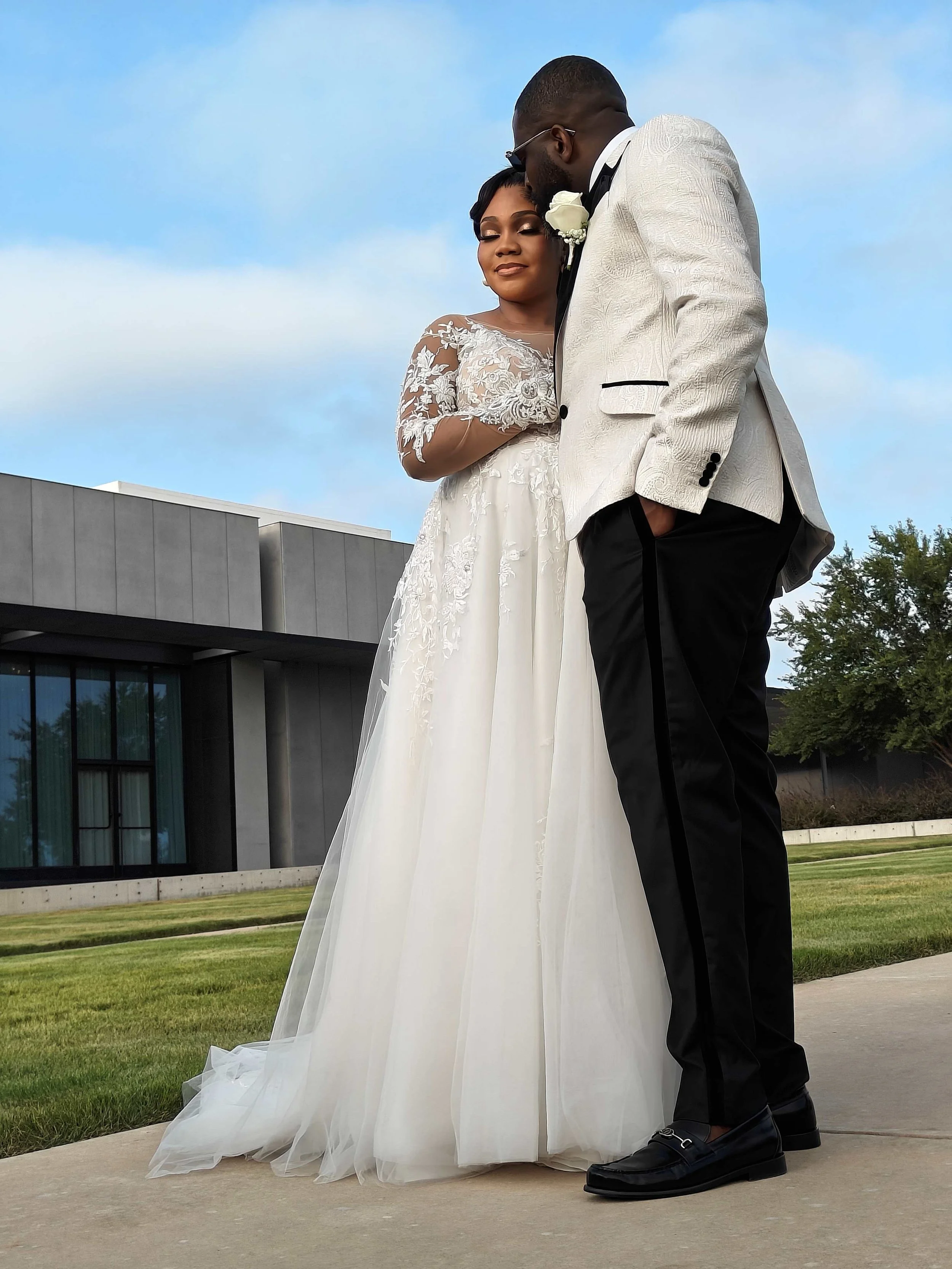 A bride in a white wedding gown with lace details and a groom in a white tuxedo jacket with black pants, standing outdoors on a concrete pathway, sharing a tender moment with the groom leaning in towards the bride's forehead.