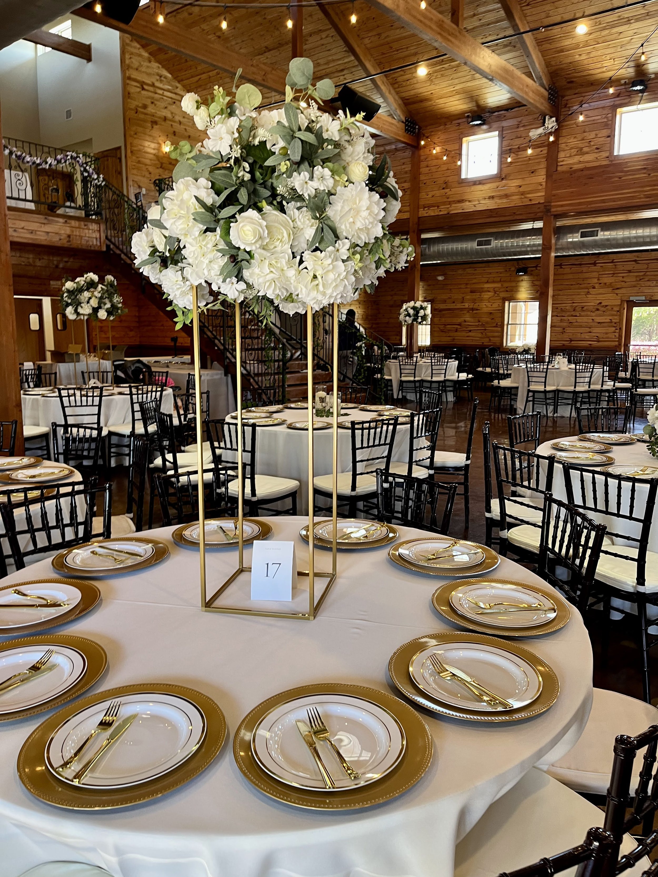 Elegant wedding reception table with a floral centerpiece of white flowers and greenery, set with gold-rimmed plates and gold utensils in a rustic wooden venue.
