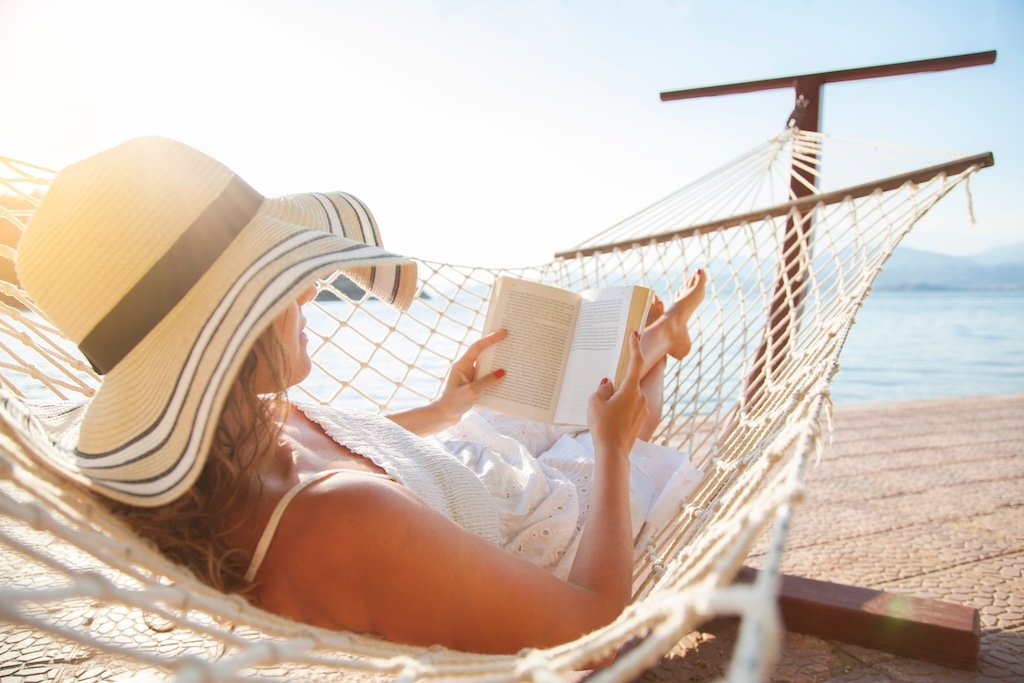 Woman relaxing on a hammock by the water, wearing a wide-brimmed hat and reading a book during the daytime.