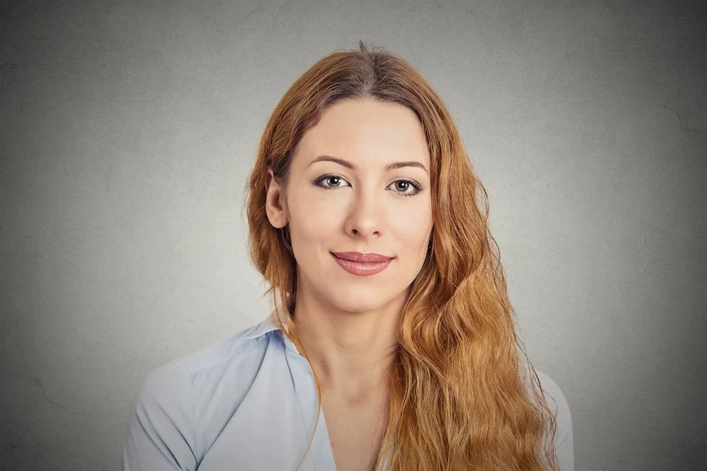 A woman with long wavy red hair, fair skin, light makeup, wearing a light blue blouse, looking at the camera with a slight smile, against a neutral gray background.