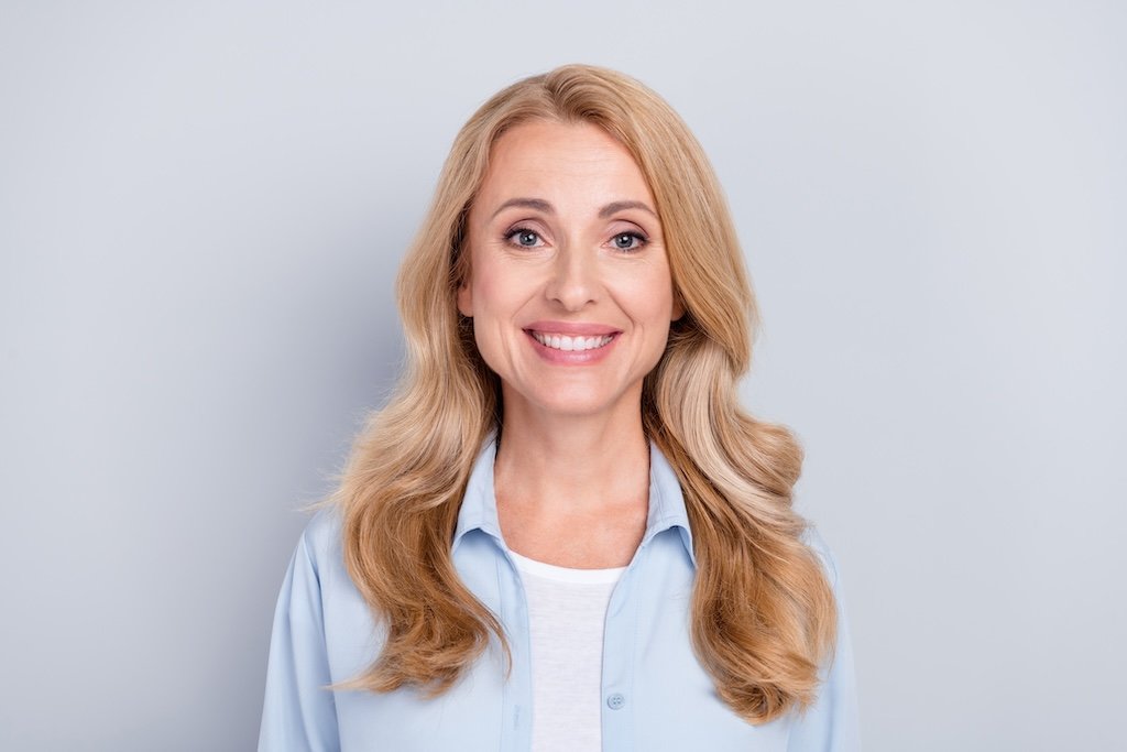 A smiling woman with long wavy blonde hair, wearing a light blue shirt, standing against a plain light gray background.