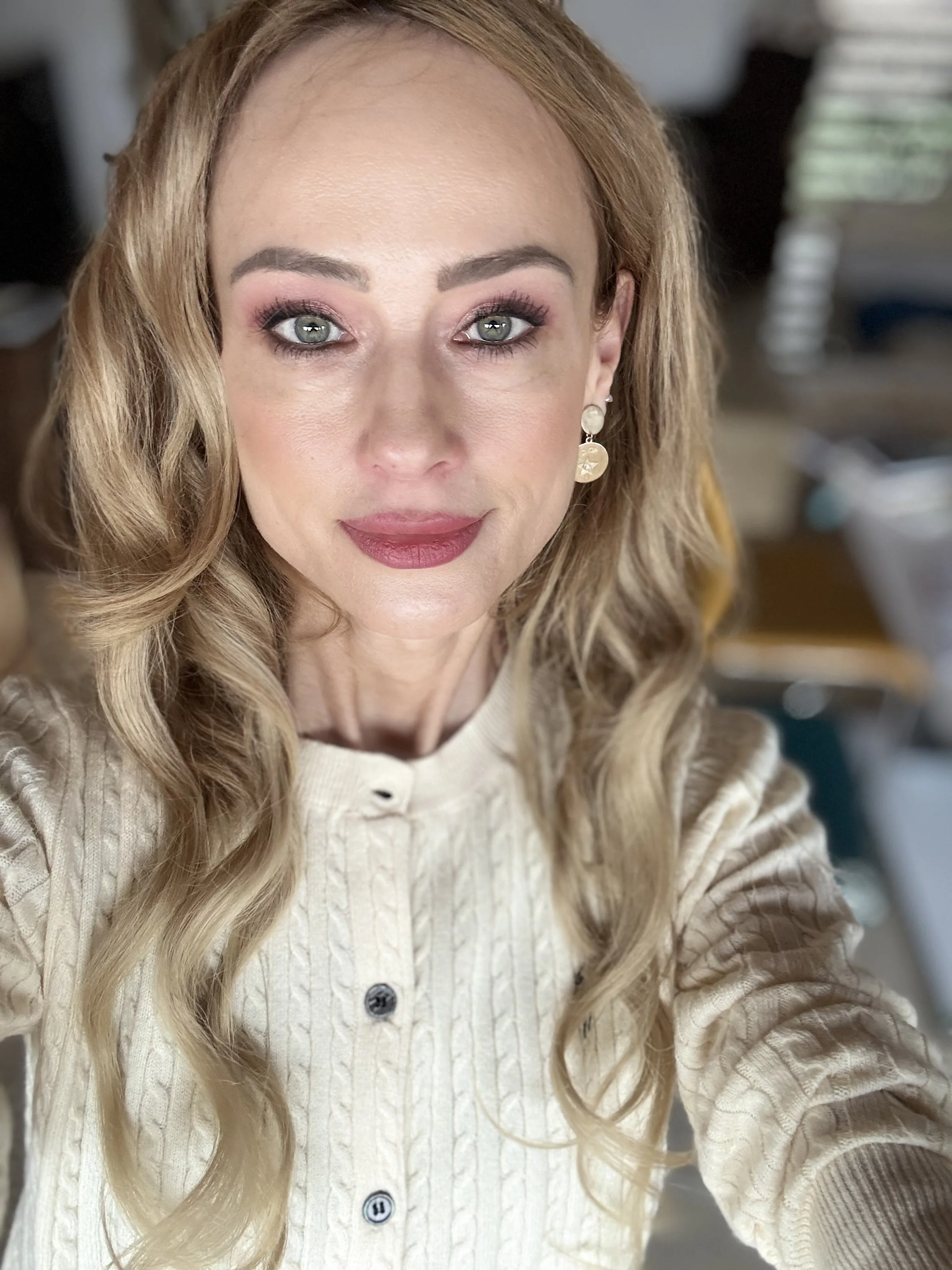 A woman with light skin, long wavy blond hair, and green eyes taking a selfie in a room with blurred background. She is wearing makeup, earrings, and a cream-colored buttoned knit sweater.