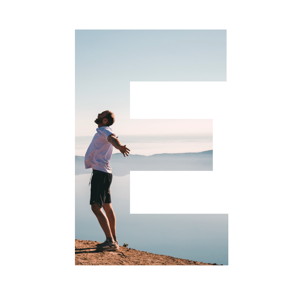 A man standing on a sandy area with his arms outstretched and head tilted back, near a body of calm water with hills in the distance during a clear day.