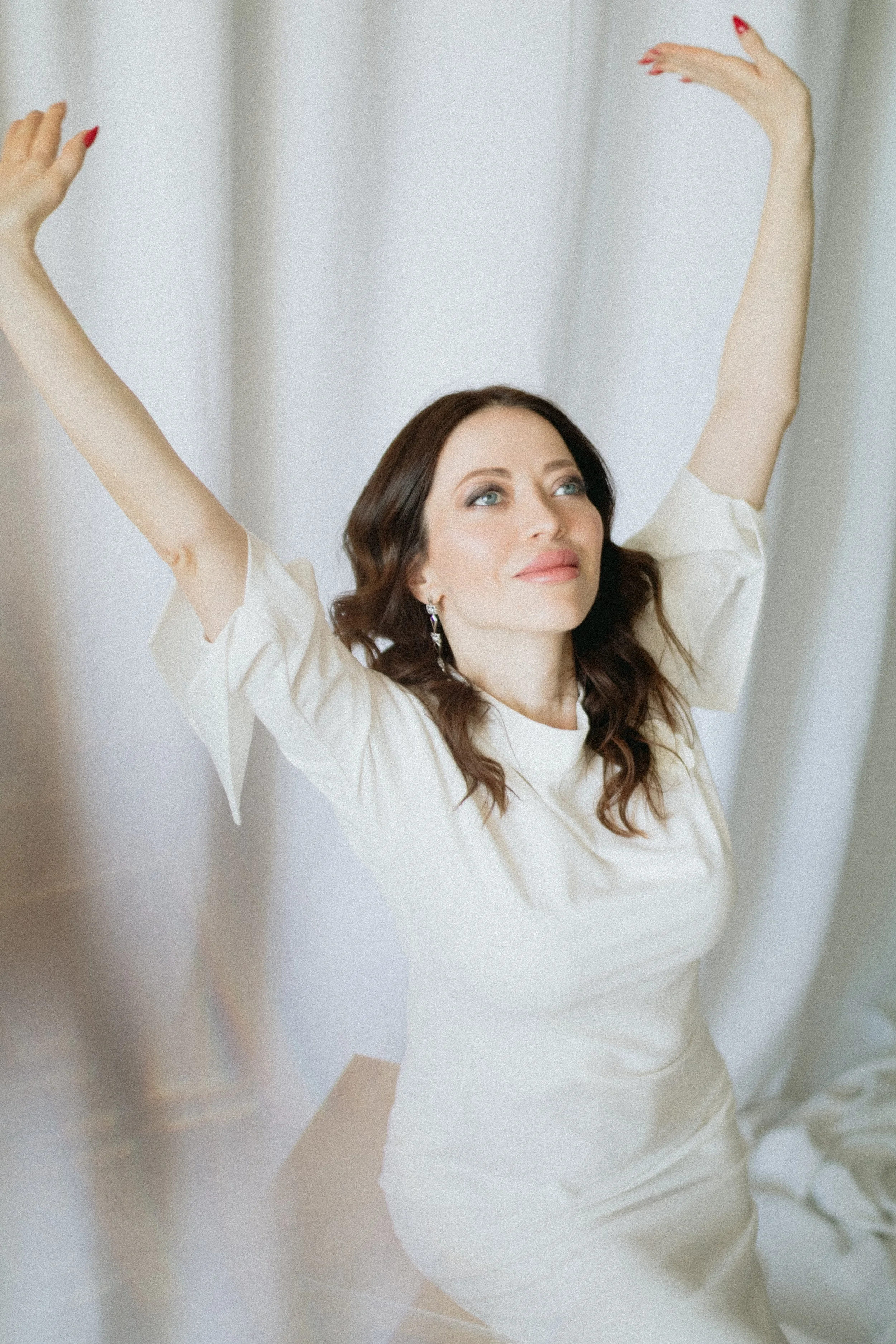 A woman with dark brown hair and blue eyes wears a cream-colored dress with puffed sleeves, raising her arms above her head with a gentle smile, standing against a soft background.