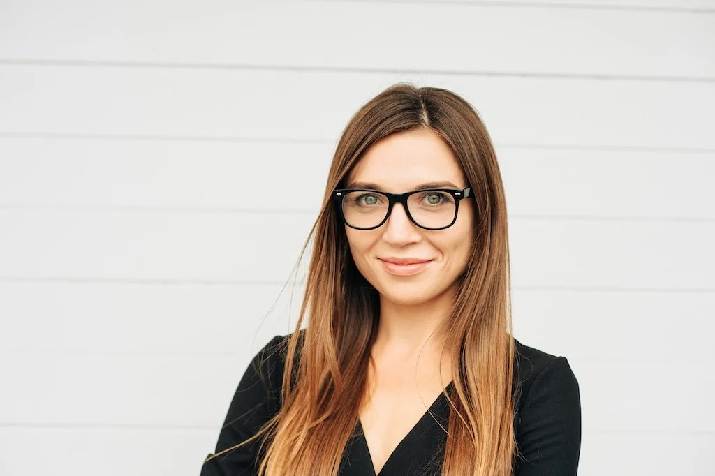 A woman with long brown hair and black glasses smiling in front of a white wall.