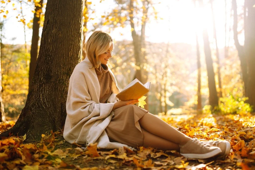 A woman reading a book while sitting on the ground against a tree in a sunlit autumn forest, surrounded by fallen leaves.