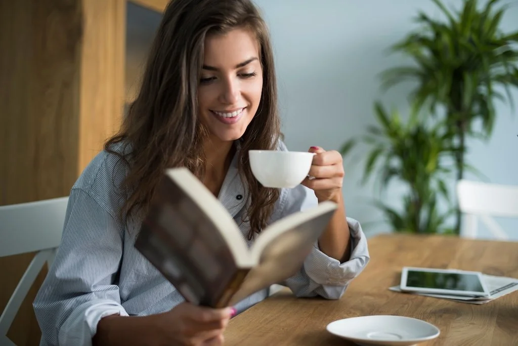 A young woman with long brown hair smiling while reading a book and drinking from a white cup at a wooden table.