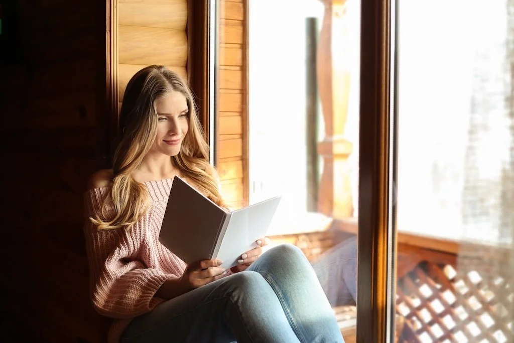 Young woman sitting by a window, reading a book, with sunlight shining inside.