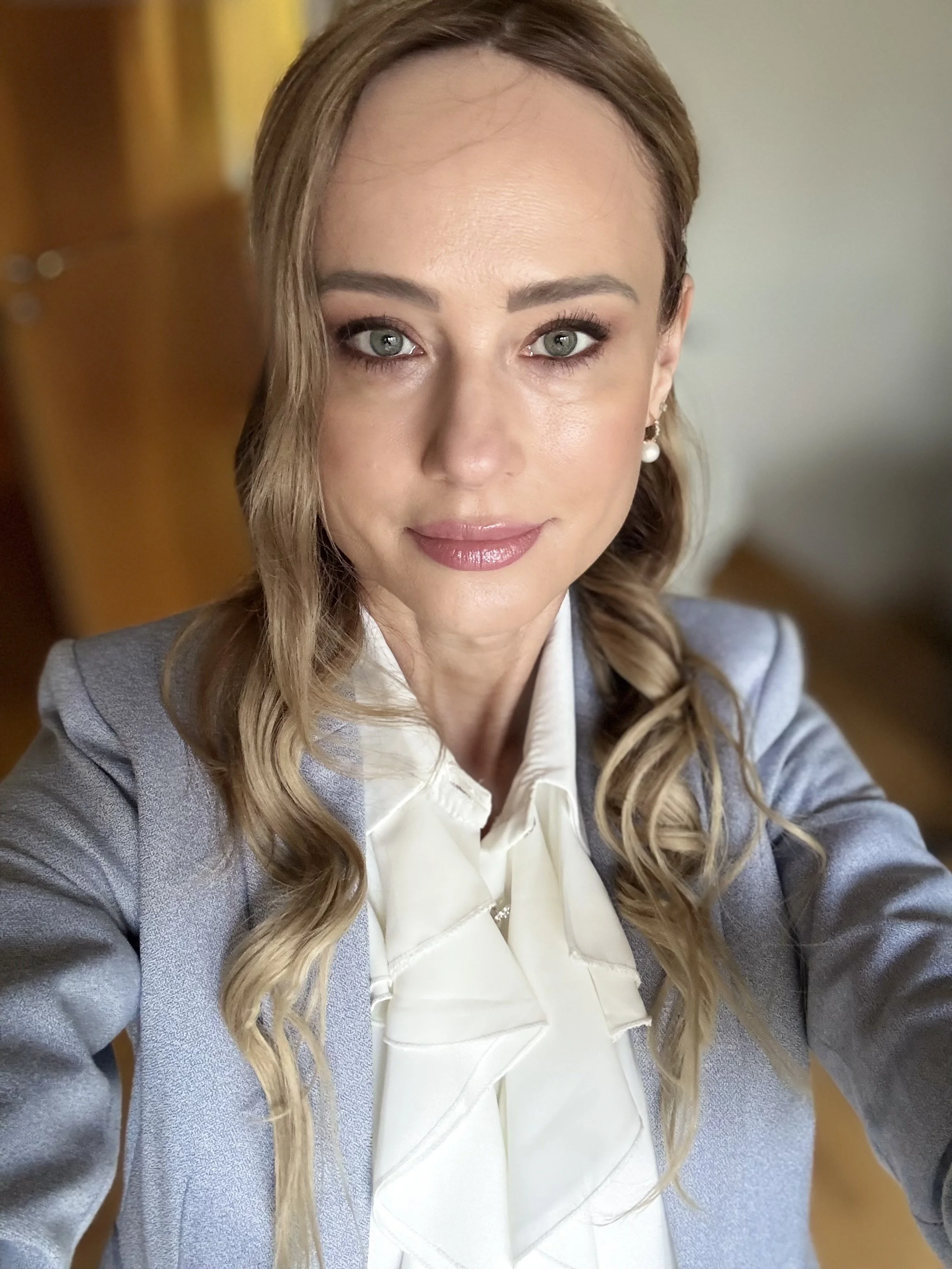 A woman with light brown hair, blue eyes, and a light complexion, dressed in professional attire, taking a selfie indoors with blurred background.