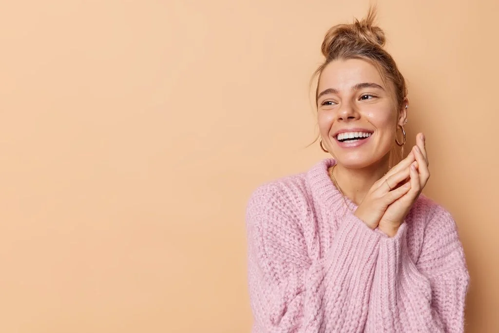 A young woman with blonde hair in a messy bun, smiling and wearing hoop earrings and a pink knitted sweater, standing against a beige background.