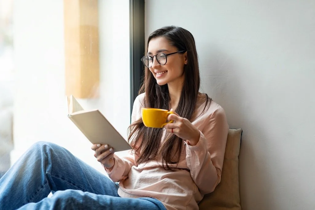 A woman with glasses smiling and reading a book while holding a yellow mug, sitting by a window.