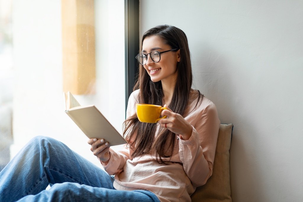 A young woman with long dark hair, glasses, and a light pink blouse is sitting by a window, smiling while reading a book and holding a yellow mug.