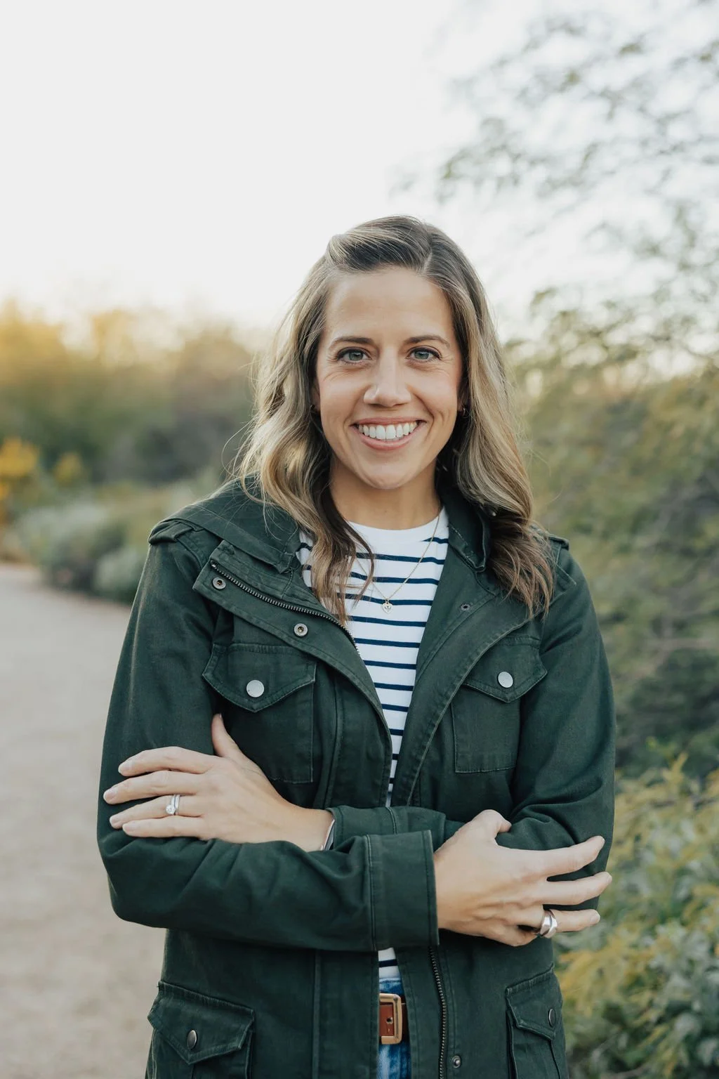 A smiling woman with brown wavy hair wearing a green jacket over a striped shirt, standing outdoors on a pathway with trees and foliage in the background.