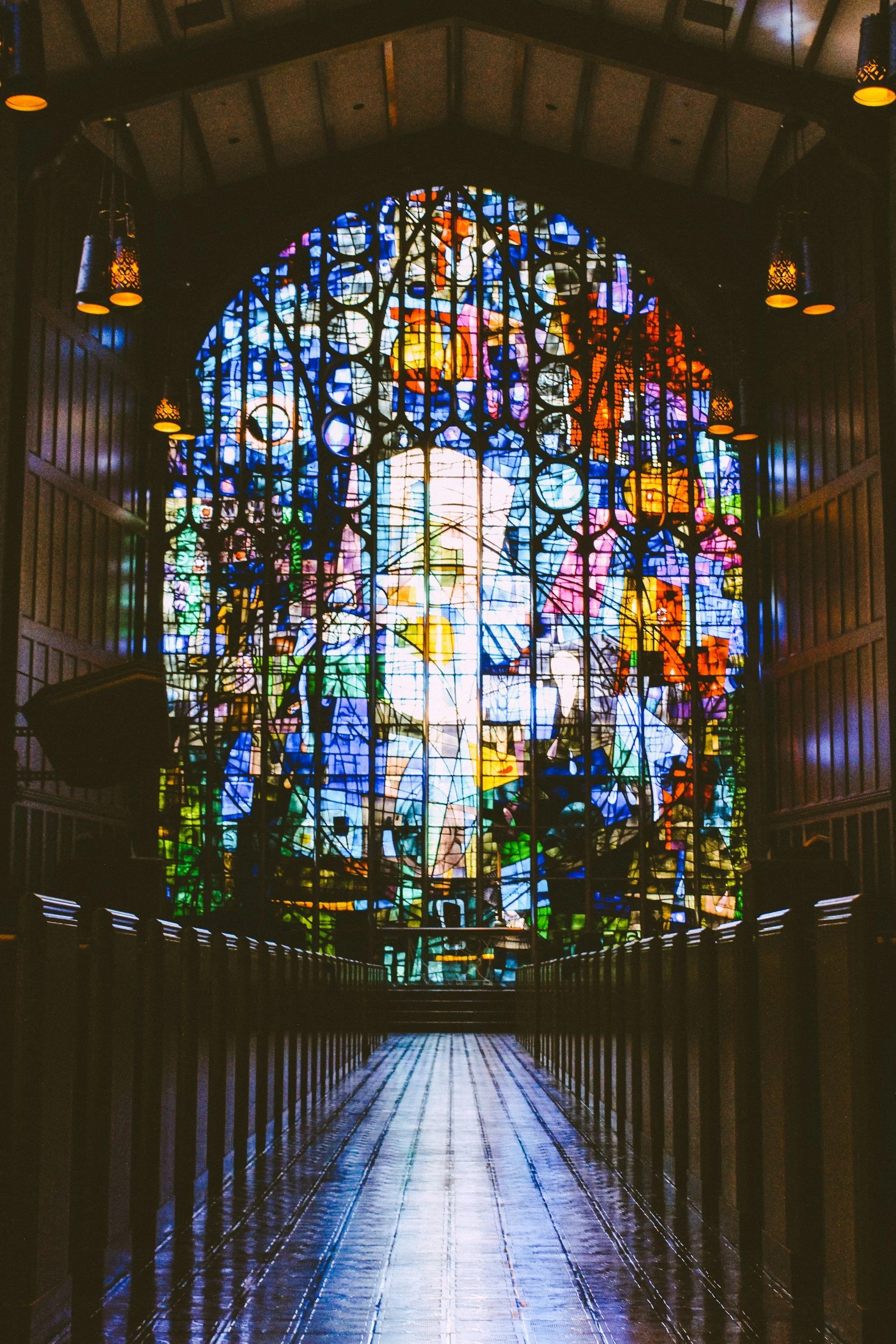 A stained glass window in a church or cathedral, with colorful abstract designs, seen from a dimly lit walkway with wooden railings on each side.