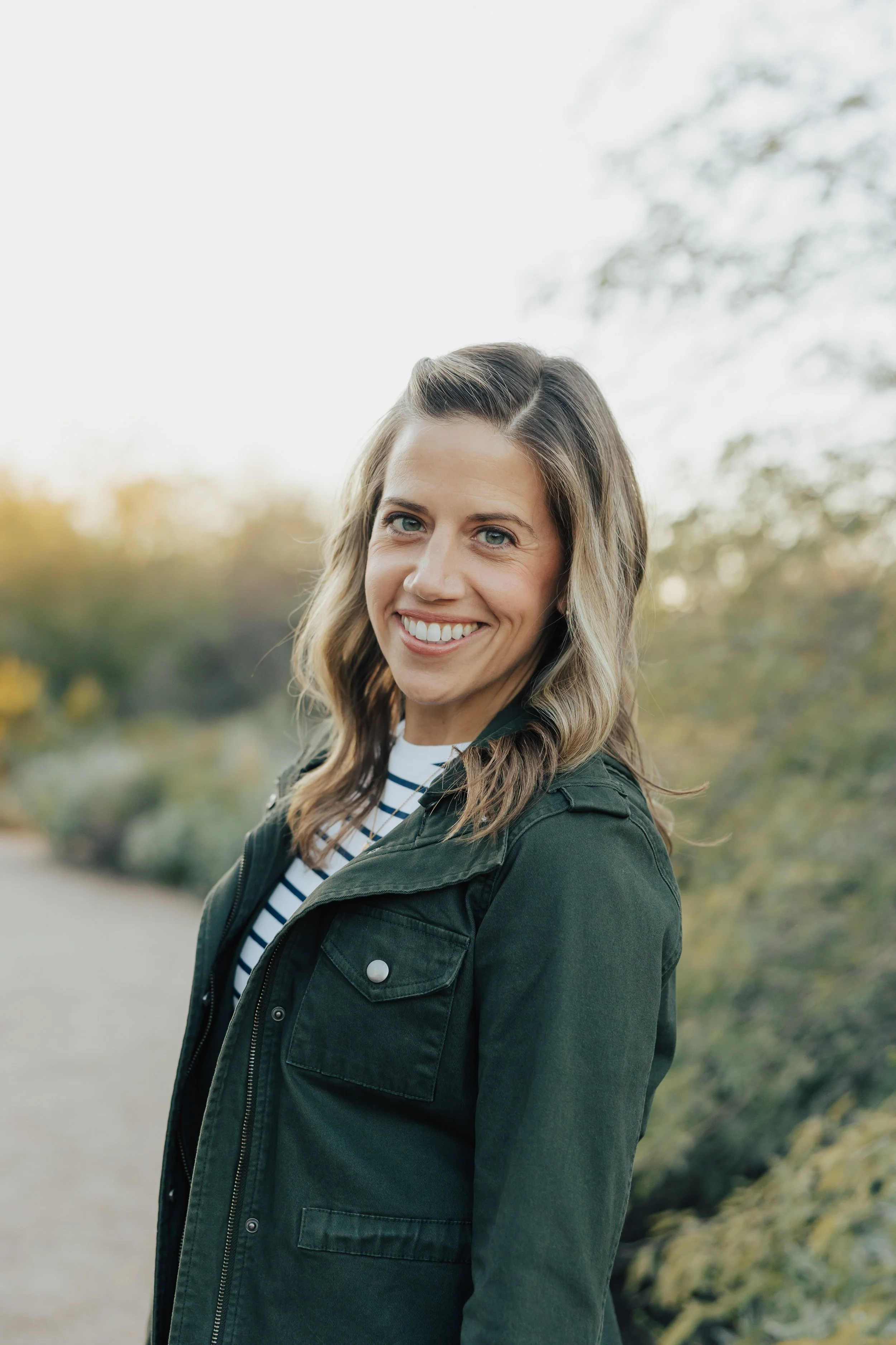 A woman with wavy light brown hair and blue eyes smiling outdoors during daytime. She is wearing a dark green jacket over a striped shirt, with blurred trees and sky in the background.